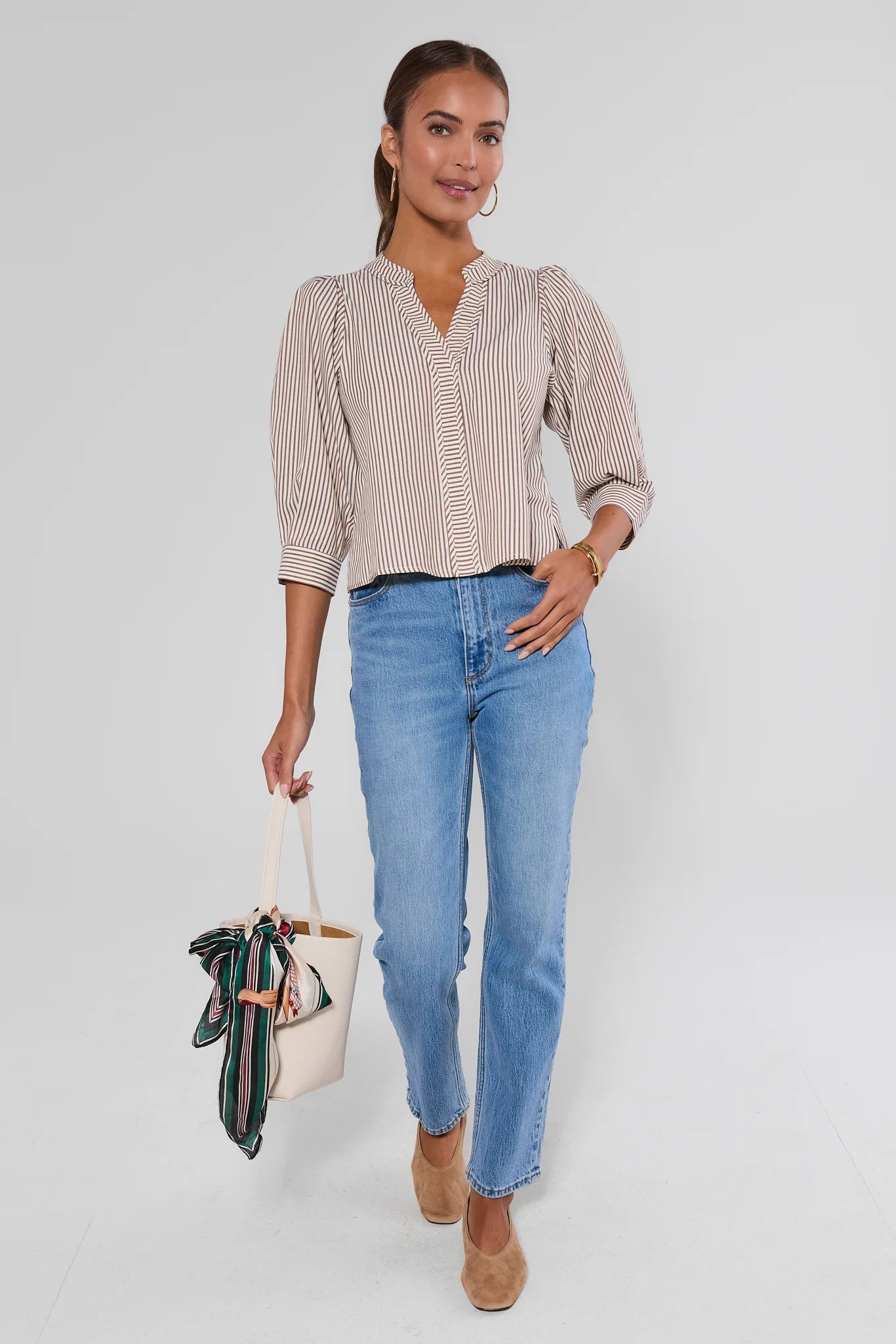 A woman modeling a beige and white vertically striped long-sleeve blouse with a split neckline, paired with light-wash blue jeans, tan shoes, and holding a cream tote bag with a green patterned scarf, against a light gray studio background.