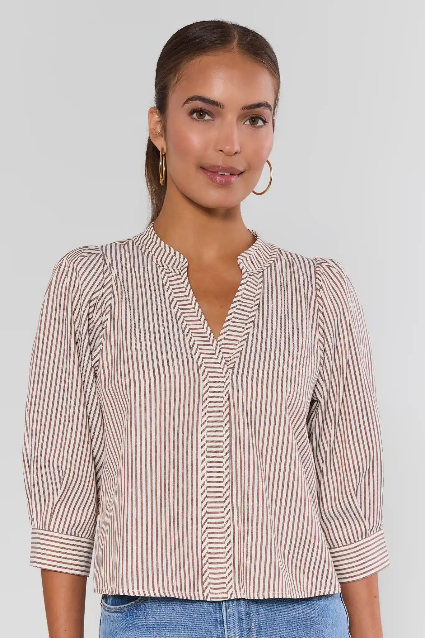 A woman wearing a beige and white vertically striped blouse with three-quarter sleeves and a split V-neckline, paired with blue jeans against a light gray studio background.