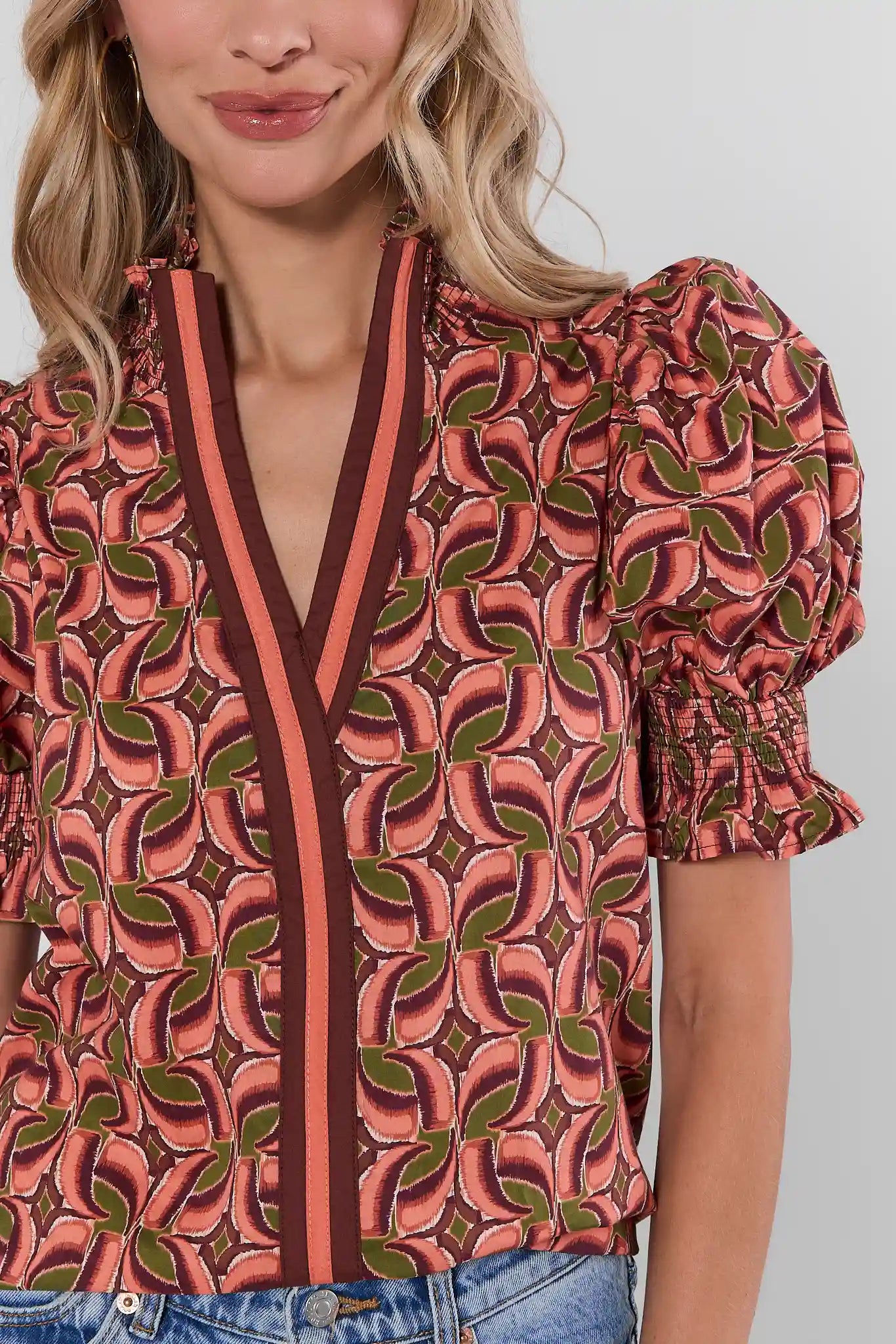 A close-up of a woman wearing a short-sleeve blouse with puff sleeves and a V-neckline, featuring an abstract leaf-like pattern in coral pink, dark brown, and olive green. The blouse is paired with blue jeans against a light gray studio background.
