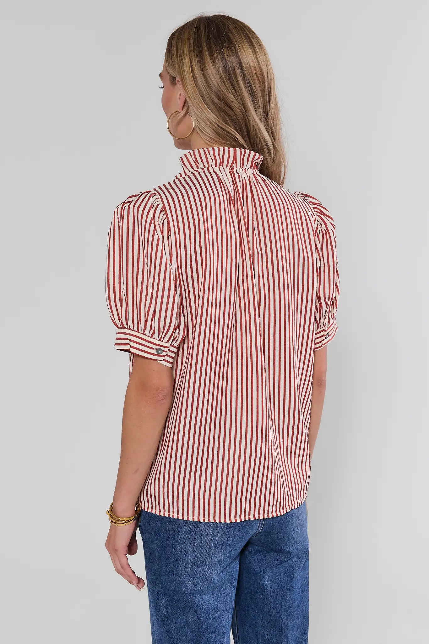 A woman shown from the back wearing a red and white vertically striped short-sleeve blouse with puff sleeves, paired with blue jeans, against a light gray studio background.