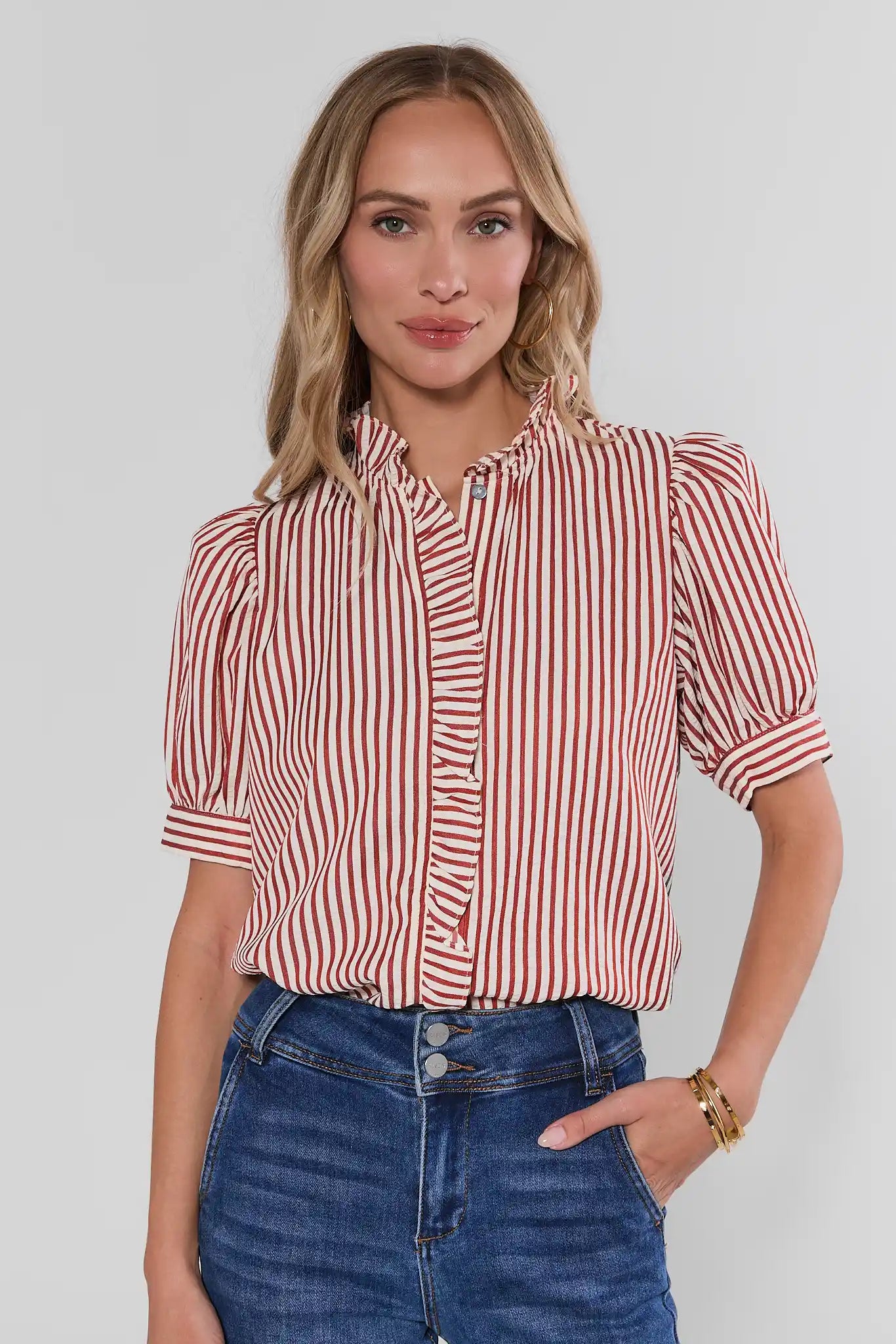 A woman wearing a red and white vertically striped short-sleeve blouse with puff sleeves and a ruffled collar, paired with blue jeans, against a light gray studio background.