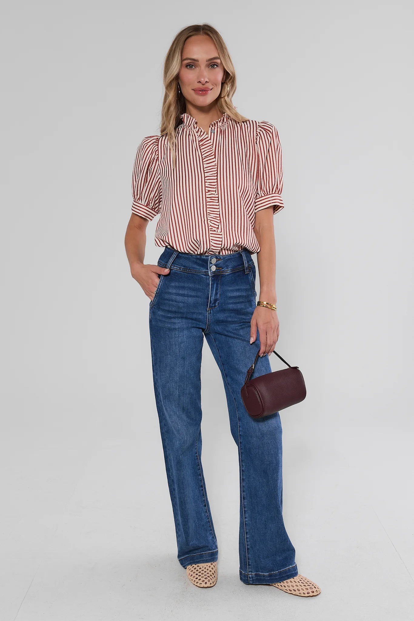 A woman modeling a short-sleeve button-up shirt with vertical red and white stripes, paired with high-waisted blue flare jeans, holding a small dark red handbag against a light gray studio background.