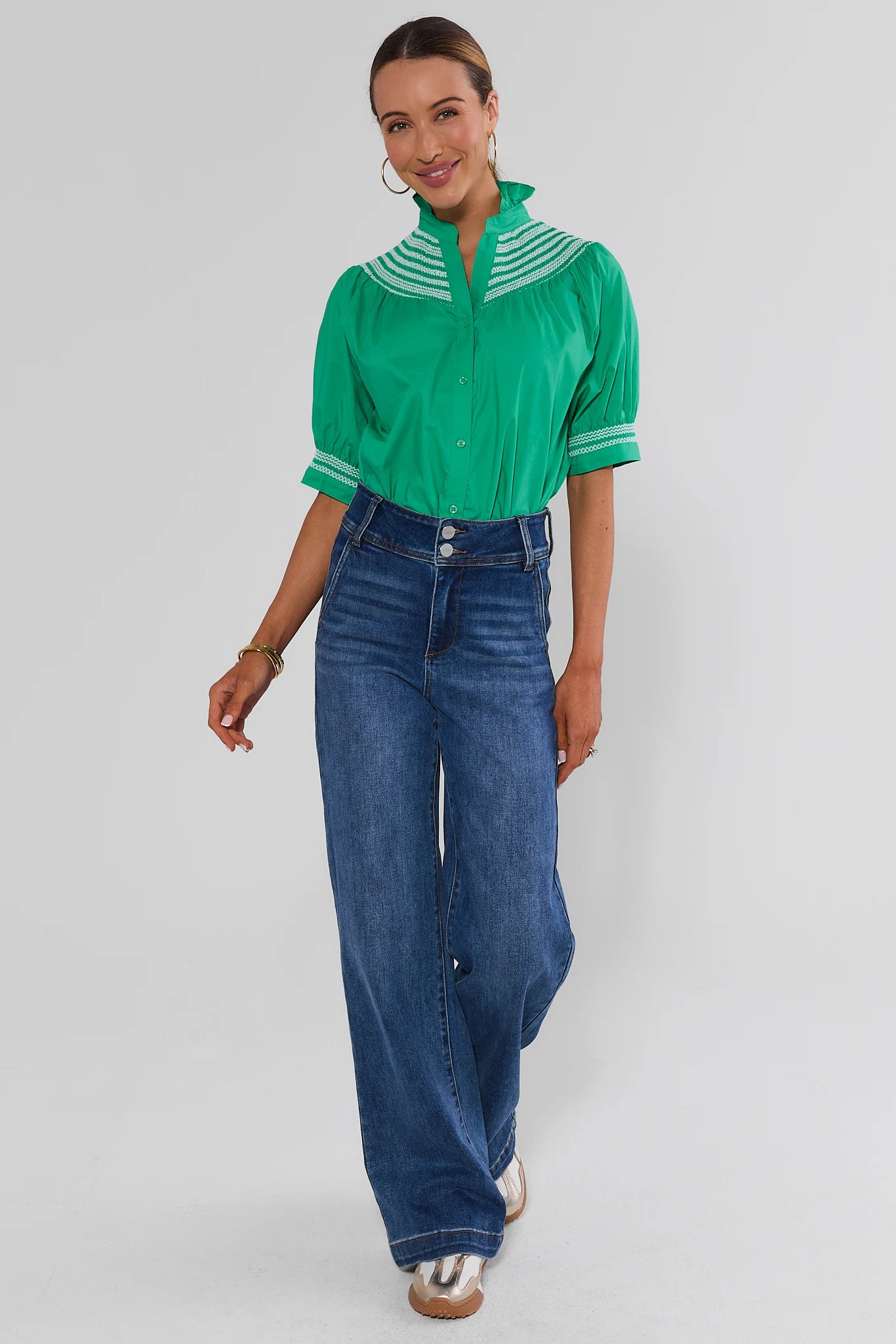 A woman modeling a bright green short-sleeve blouse with white striped collar and cuffs, paired with high-waisted wide-leg blue jeans and white slide sandals, against a light gray studio background.