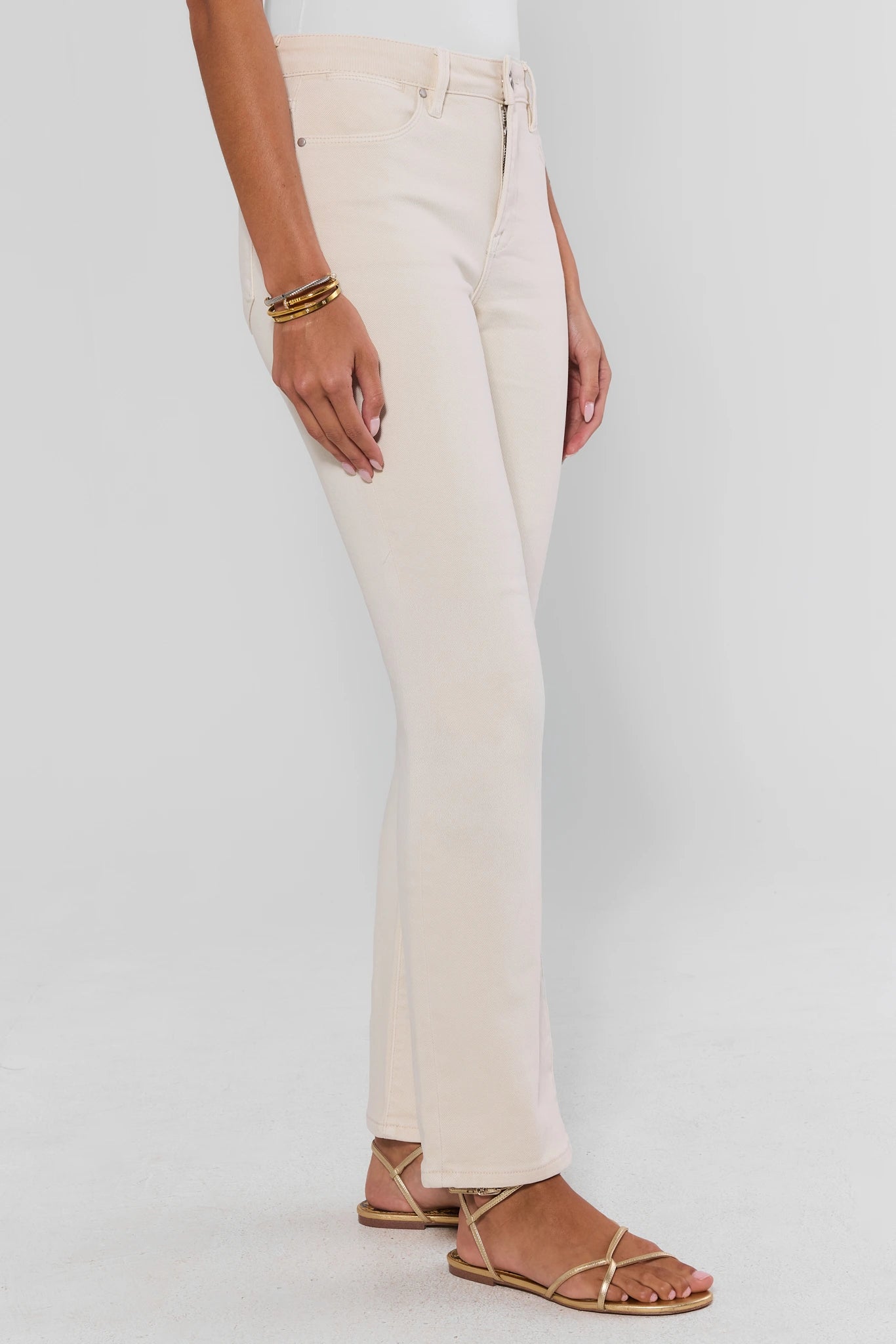 A model wearing a pair of light cream or off-white flare jeans, paired with gold strappy flat sandals and gold bracelets, shown from the waist down against a light gray studio background.