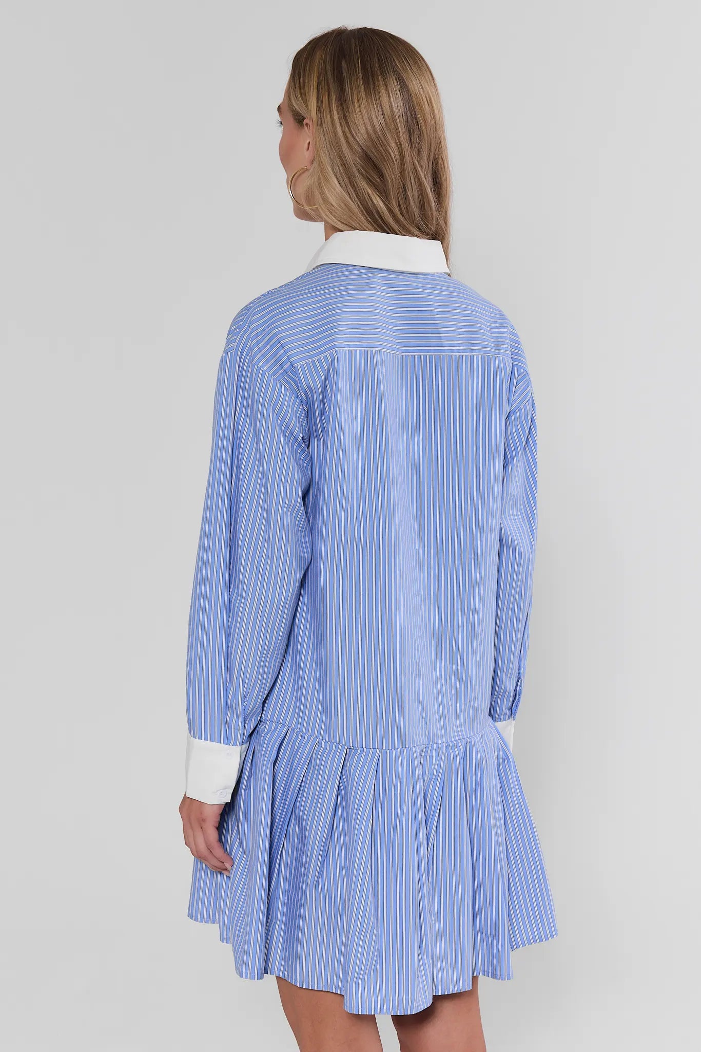 A woman shown from the back wearing a light blue and white vertically striped shirt dress with a white collar and white cuffs, set against a plain light gray studio background.