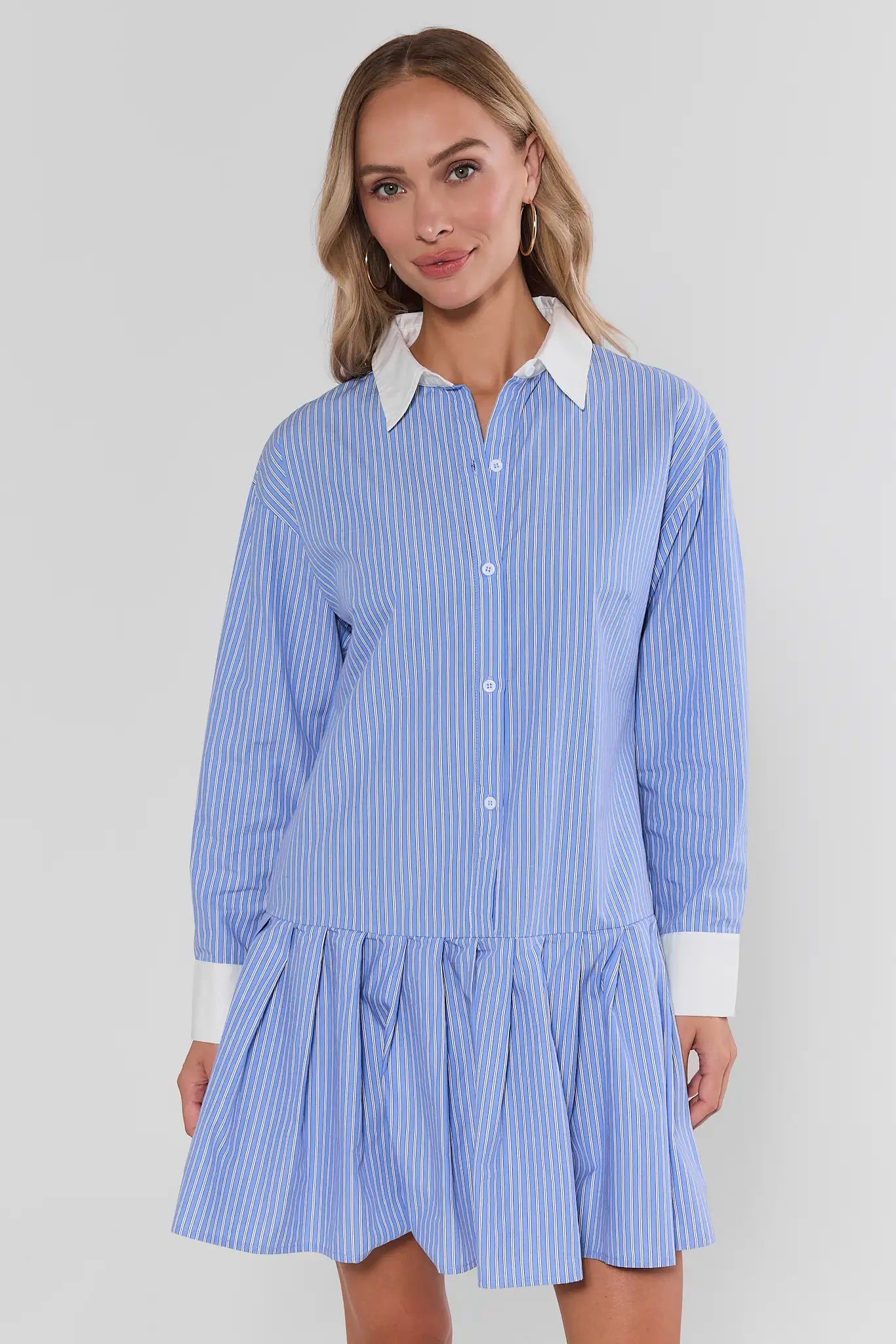 A woman modeling a light blue and white vertically striped shirt dress with a white pointed collar and white cuffs, set against a plain light gray studio background.