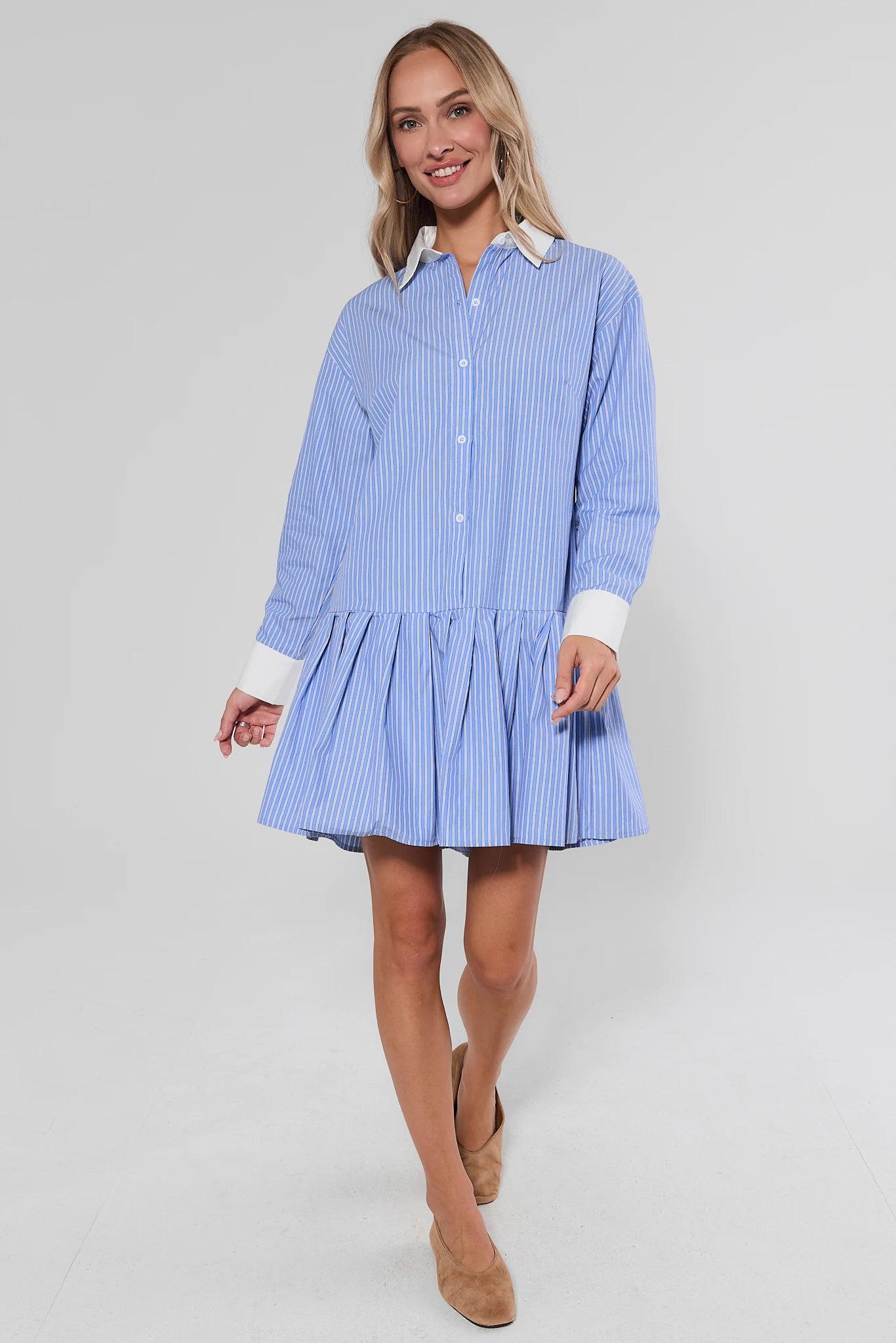 A woman modeling a light blue and white vertically striped mini shirt dress with a flared skirt, button-up front, and white contrast collar and cuffs, photographed against a plain studio background.