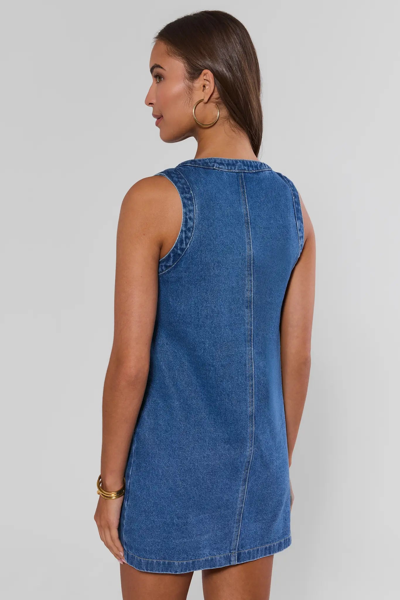 A woman shown from the back wearing a sleeveless blue denim dress against a plain light gray studio background.