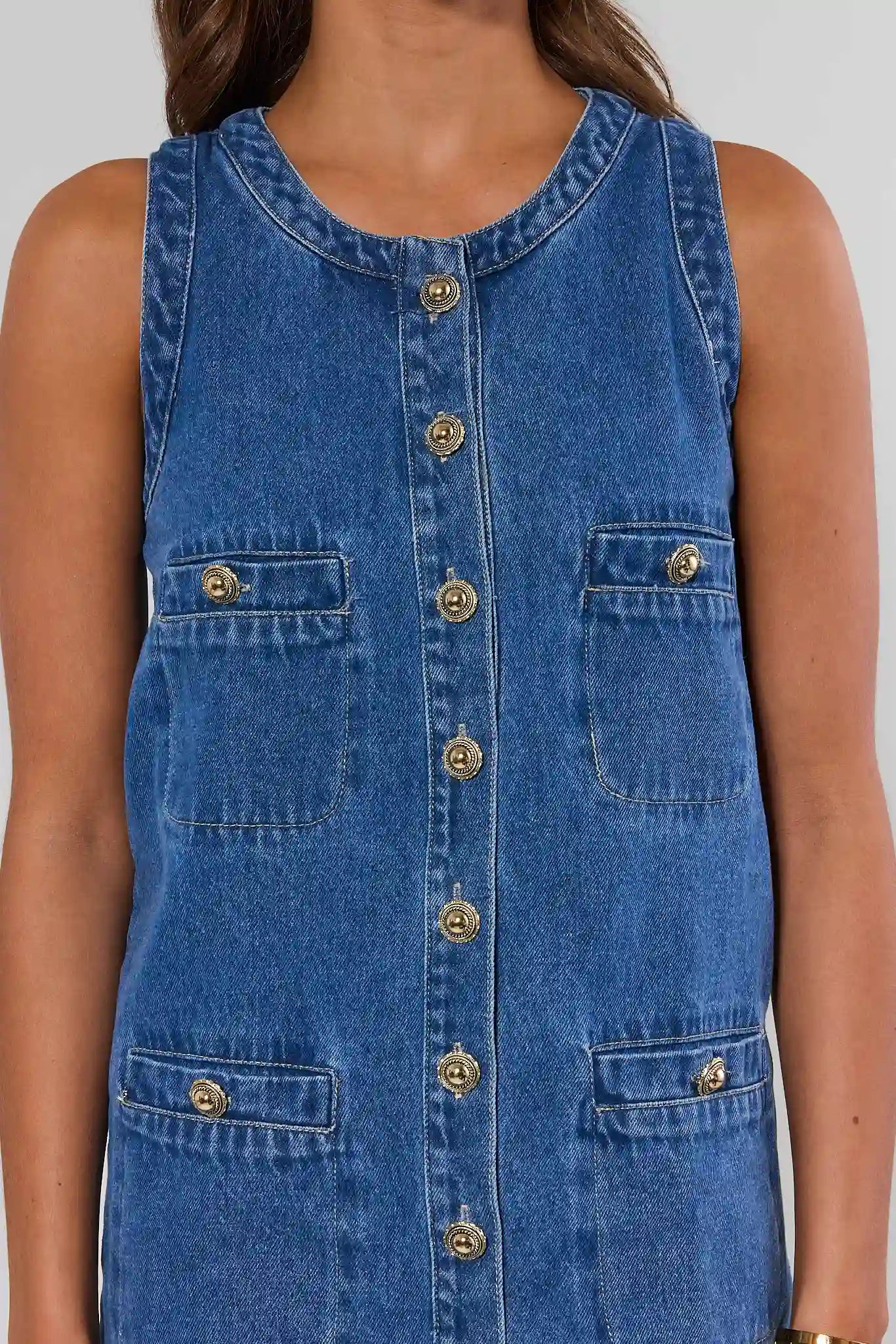 A close-up of a person wearing a sleeveless blue denim dress with gold-tone buttons and front patch pockets.