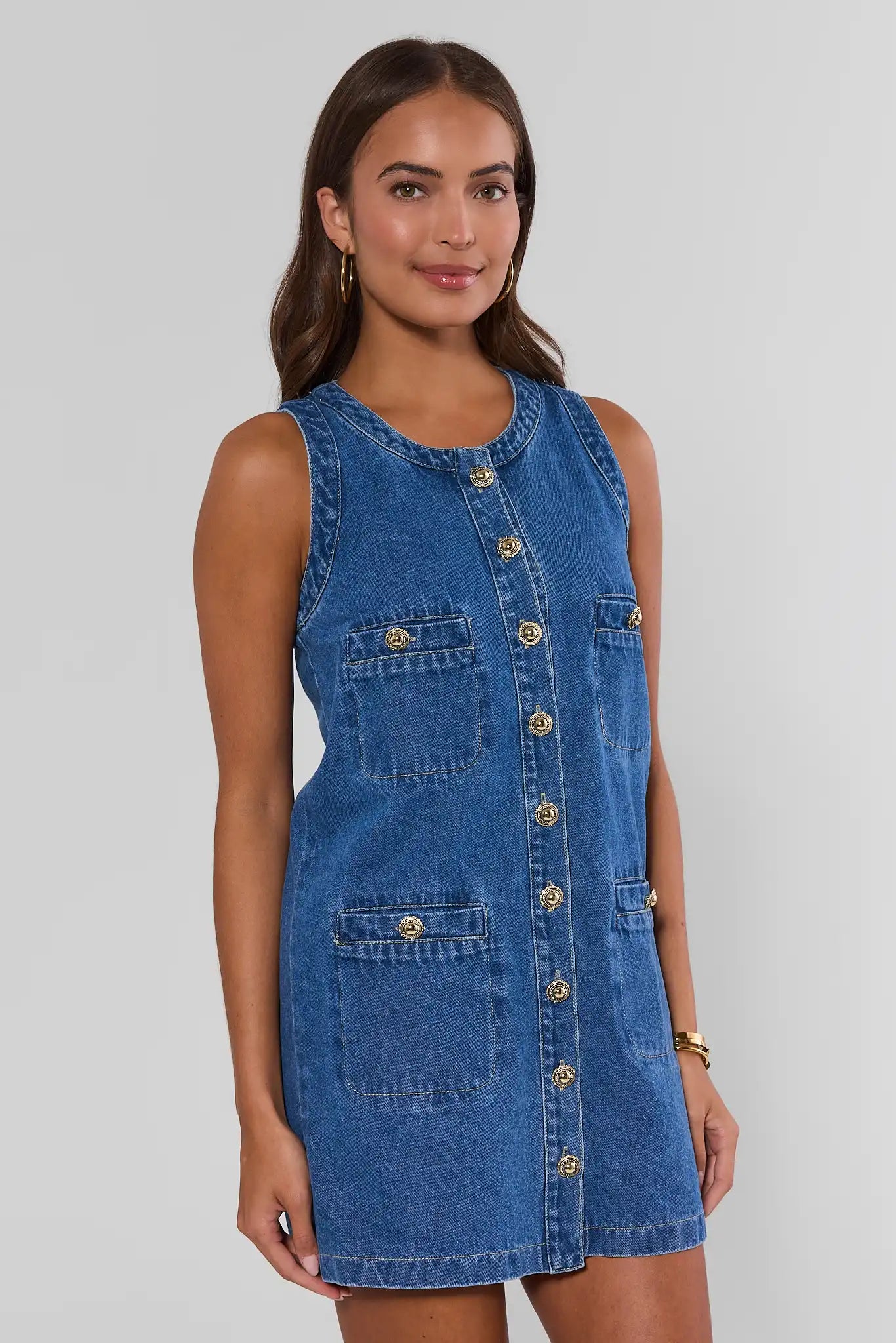 A woman modeling a sleeveless blue denim dress with gold-tone buttons down the front and on the chest pockets, photographed against a light gray studio background.