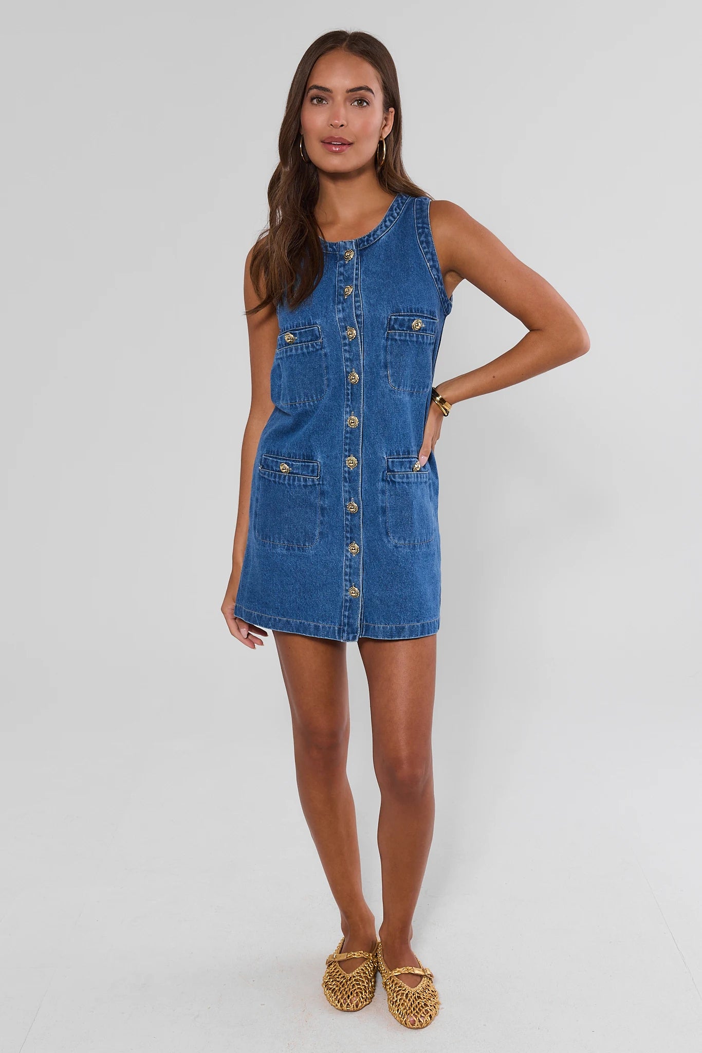 A woman modeling a sleeveless blue denim pinafore dress with a button-front closure and front pockets, paired with tan woven flat shoes, against a light gray studio background.