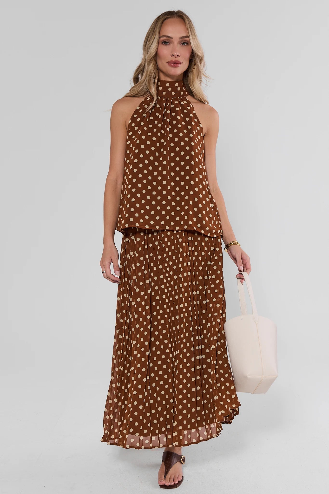 A woman modeling a brown sleeveless halter-neck maxi dress with a white polka dot pattern, paired with brown flat sandals and holding a white tote bag against a light gray studio background.