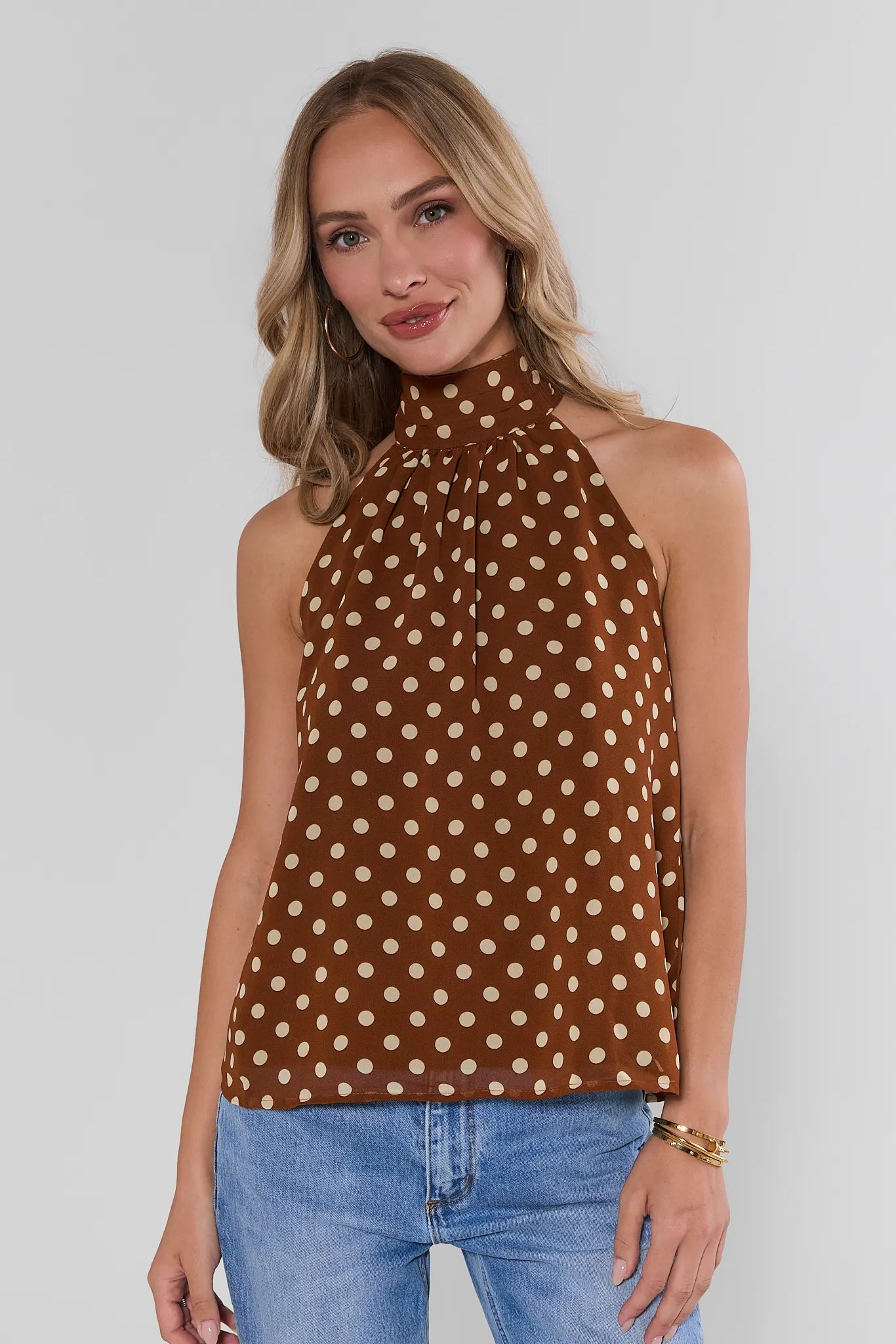 A woman wearing a brown halter-neck top with a beige polka dot pattern, paired with blue jeans, against a light gray studio background.
