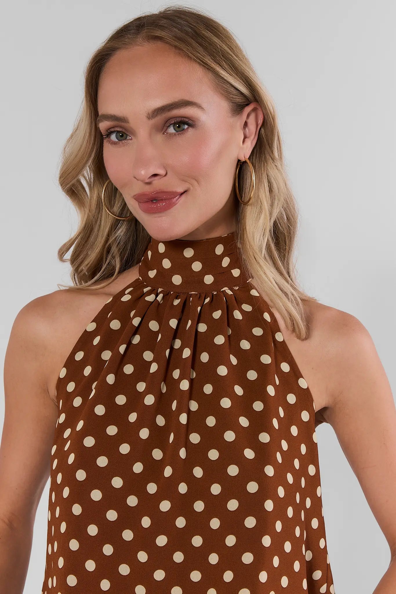 A woman wearing a brown halter-neck top with a cream polka dot pattern, shown from the chest up against a light gray studio background.
