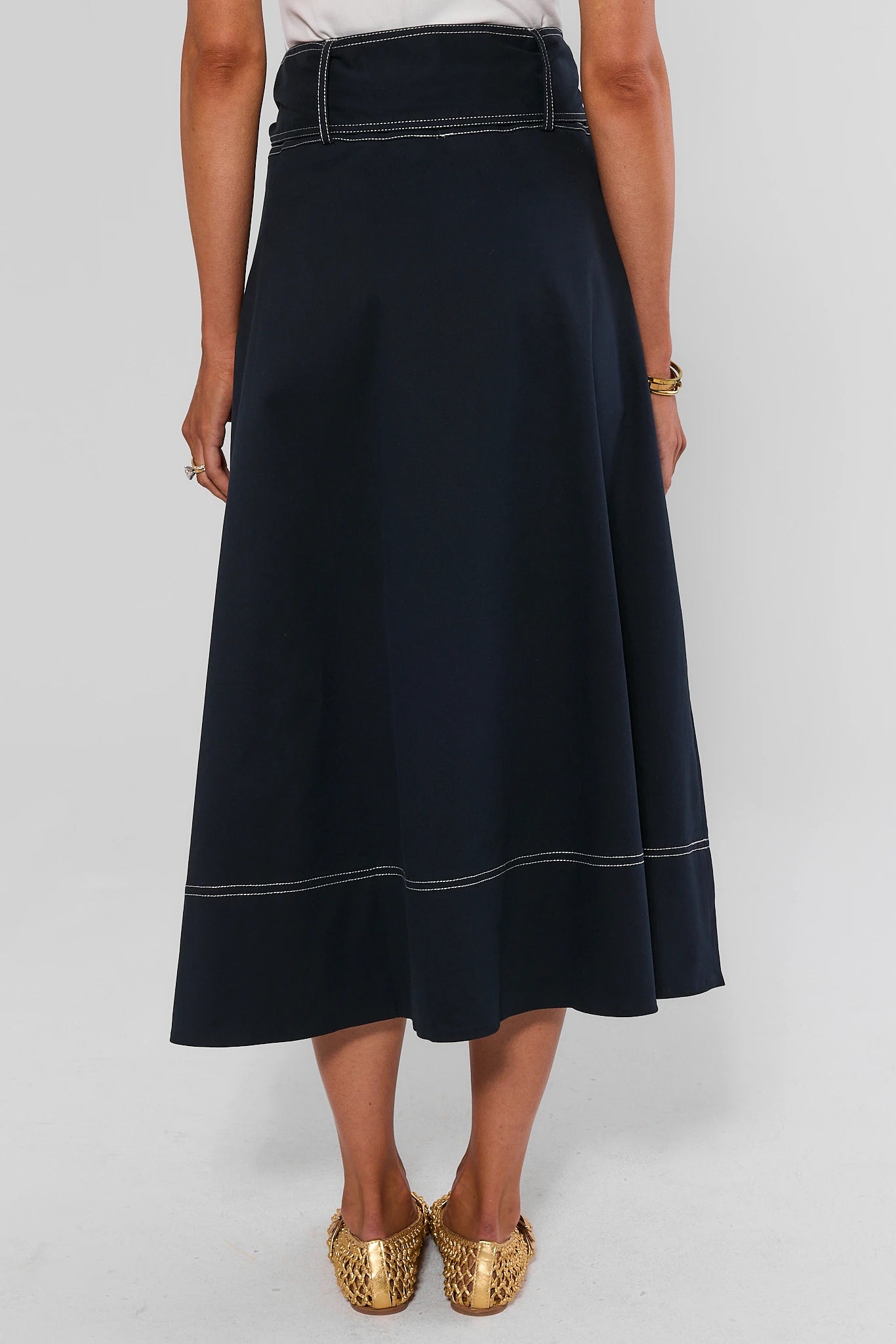 A dark navy blue midi skirt with white contrast stitching, worn by a model with gold flat shoes against a light gray background.