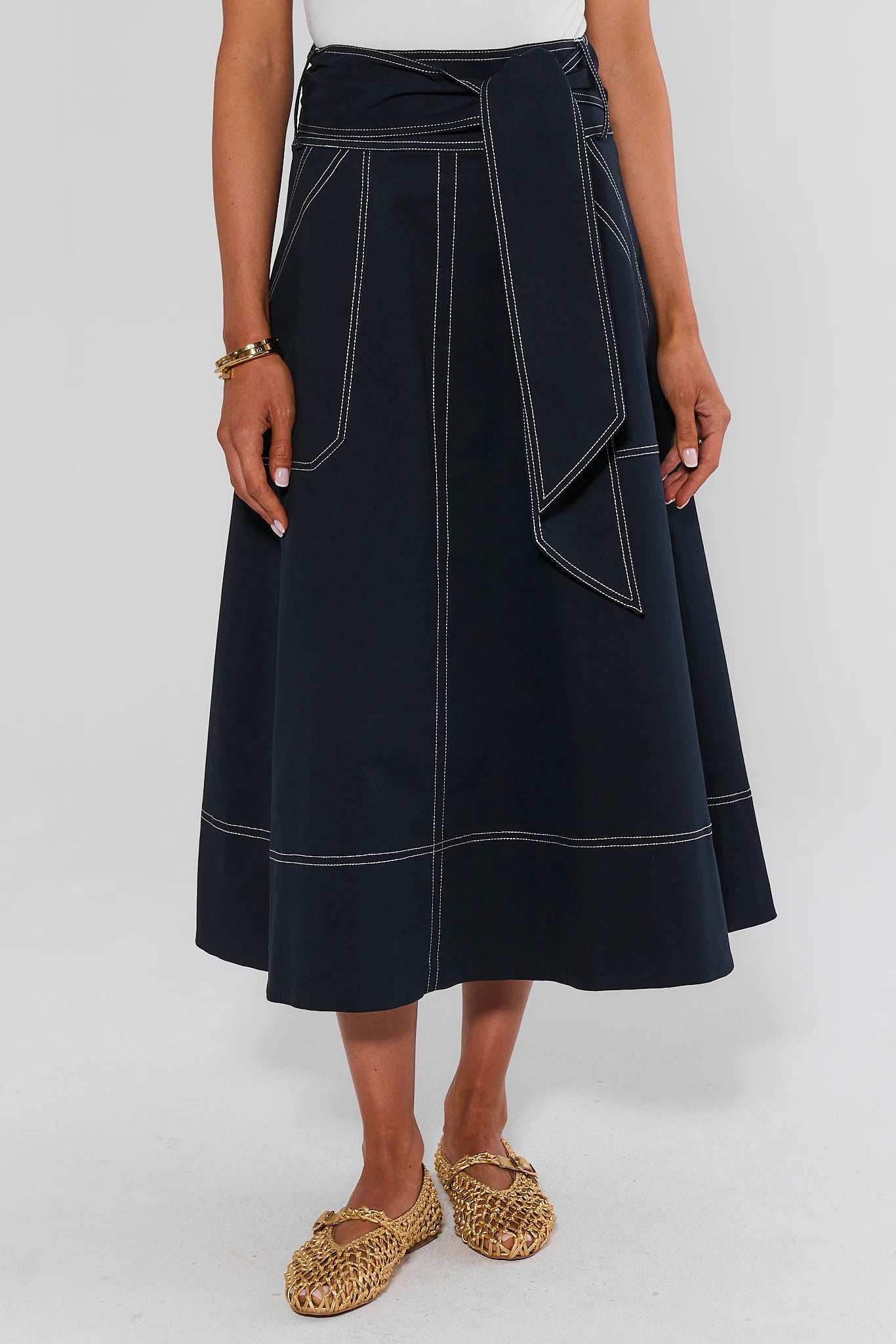 A model wearing a dark navy blue midi skirt with white contrast stitching, a matching waist tie, and large front patch pockets, paired with woven espadrille flats.