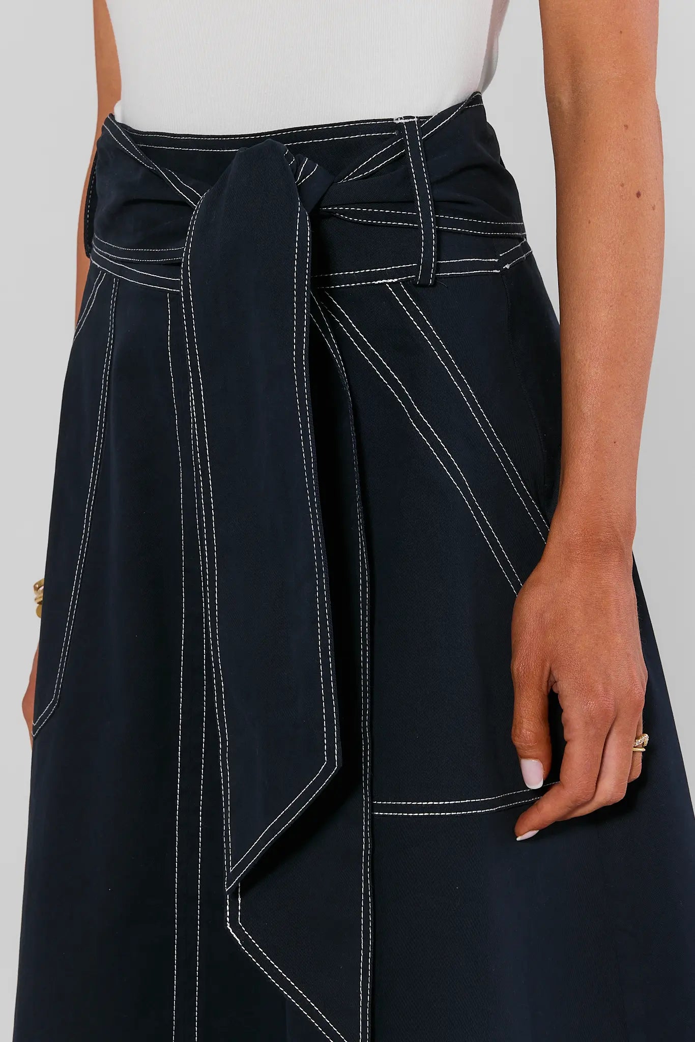 Close-up of a model wearing a dark navy wrap skirt with white contrast stitching and a tie waist, paired with a white top against a light gray background.