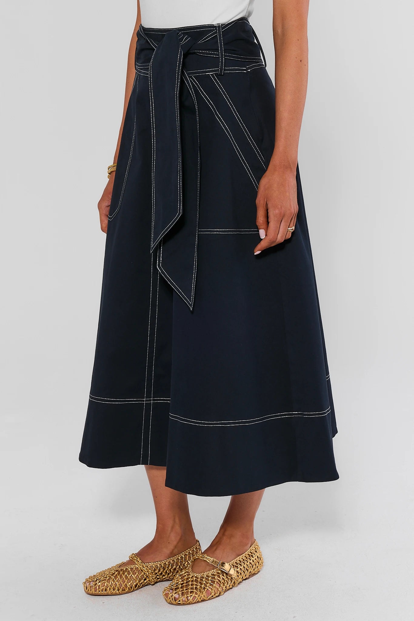 A model wearing a dark navy blue wrap midi skirt with white contrast stitching, paired with woven straw flat shoes, against a light gray studio background.