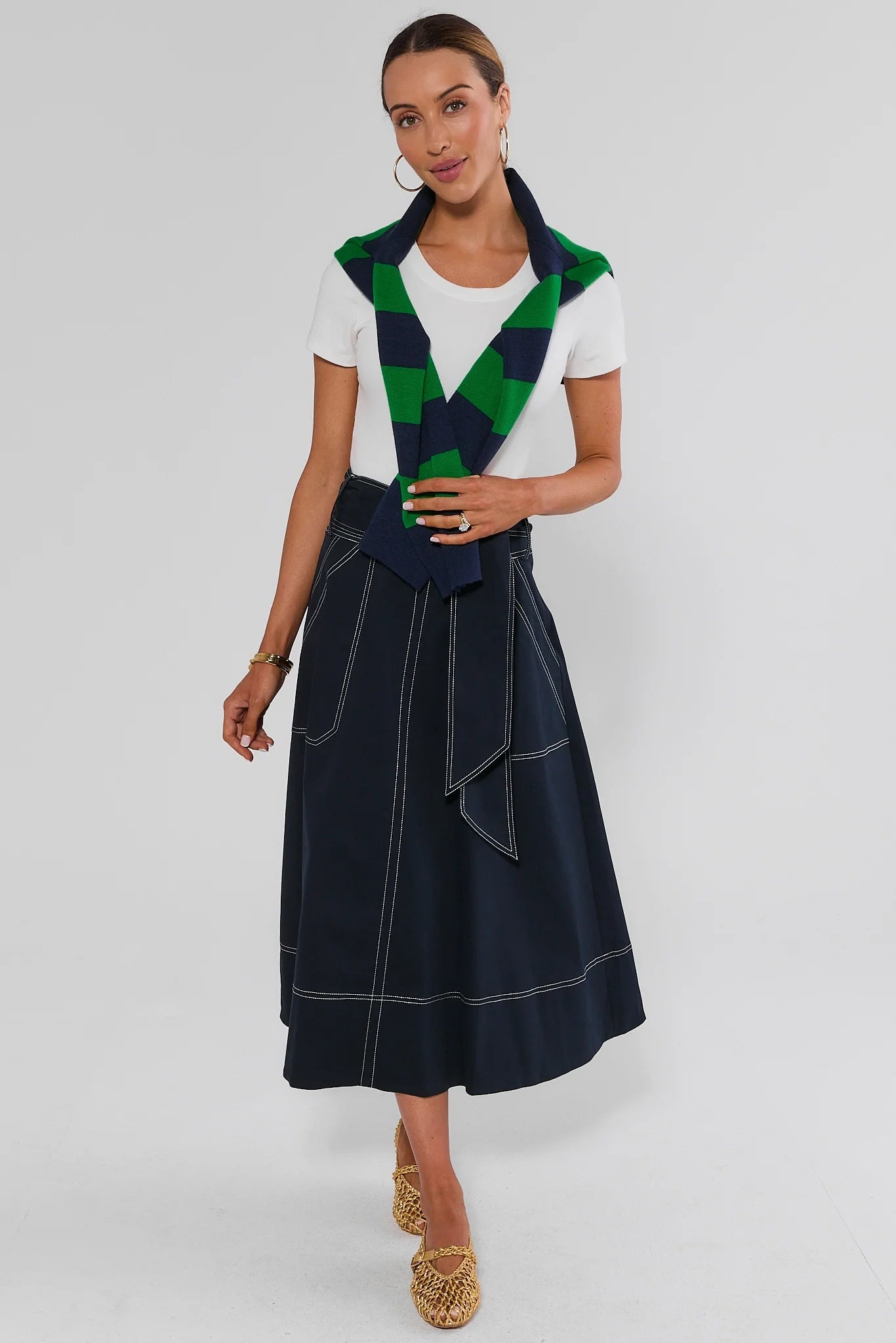 A woman models a navy blue midi skirt with white contrast stitching, styled with a white t-shirt and a green and navy checkered scarf, against a light gray studio background.
