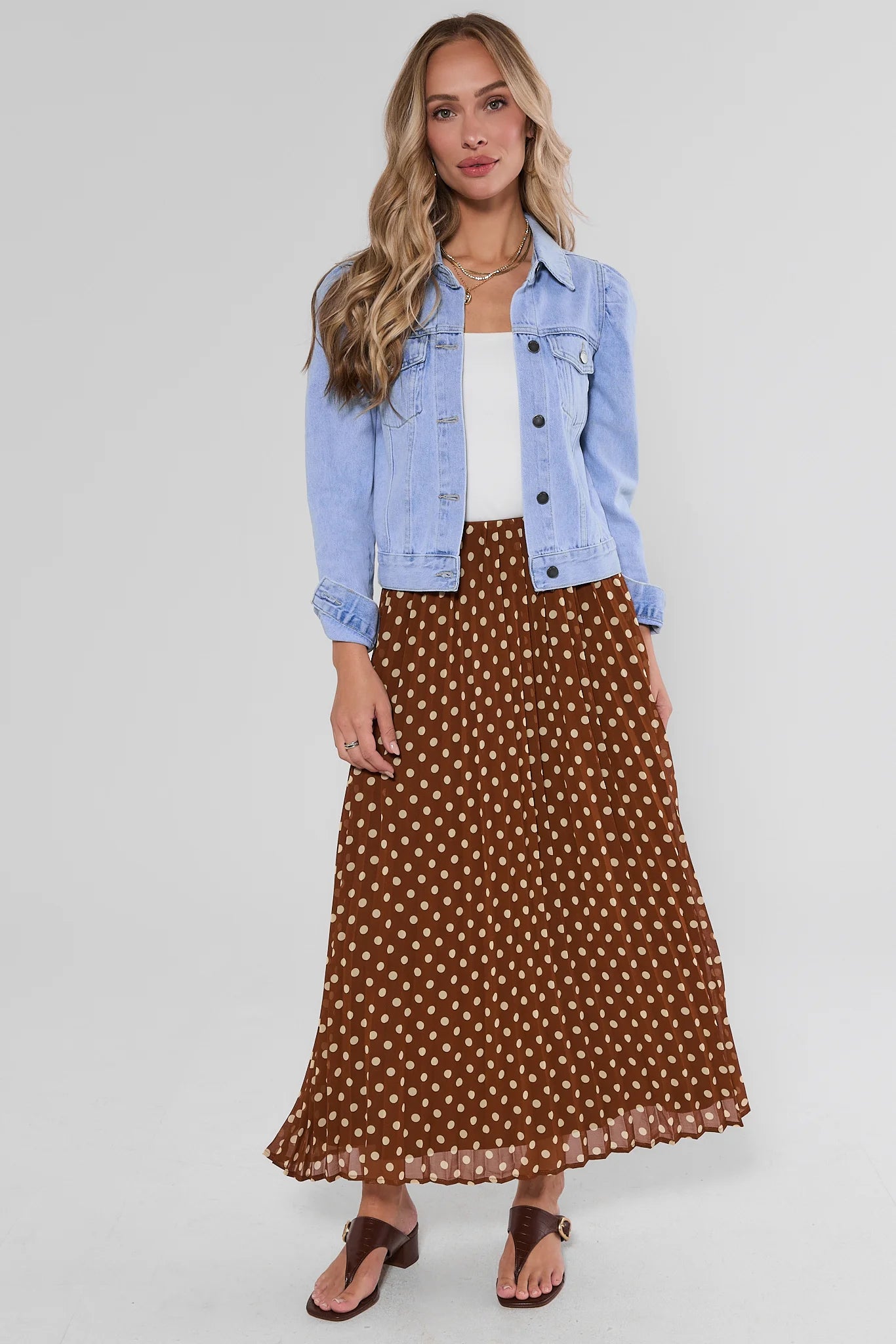 A woman modeling a brown midi skirt with a white polka dot pattern, styled with a light blue denim jacket, white top, and brown sandals against a light gray background.