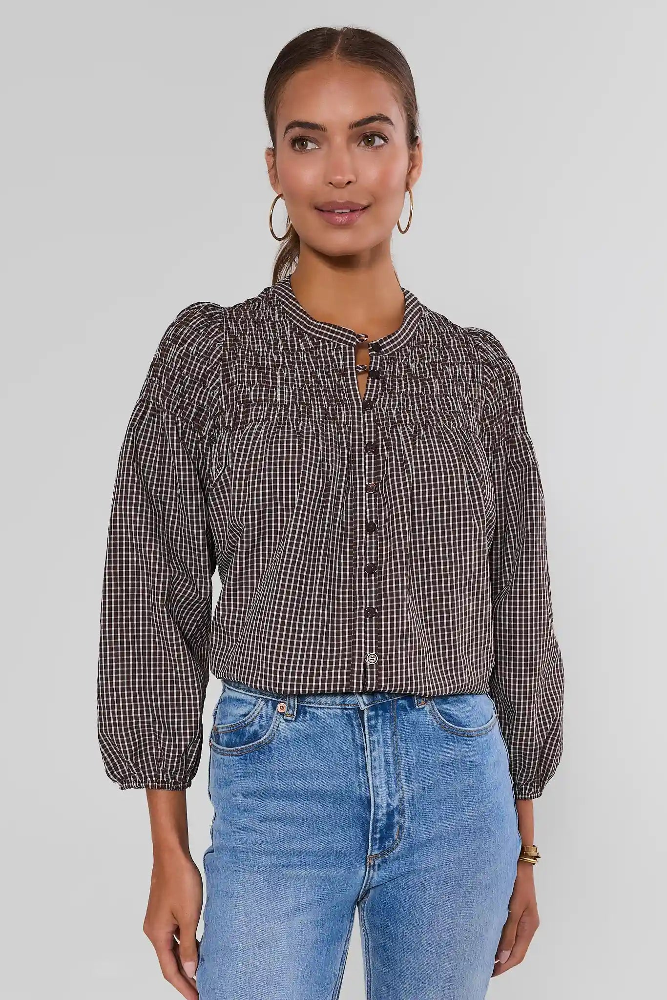 A woman wearing a brown and black gingham long-sleeve blouse with puff sleeves and a smocked yoke, paired with light blue jeans, against a light gray studio background.