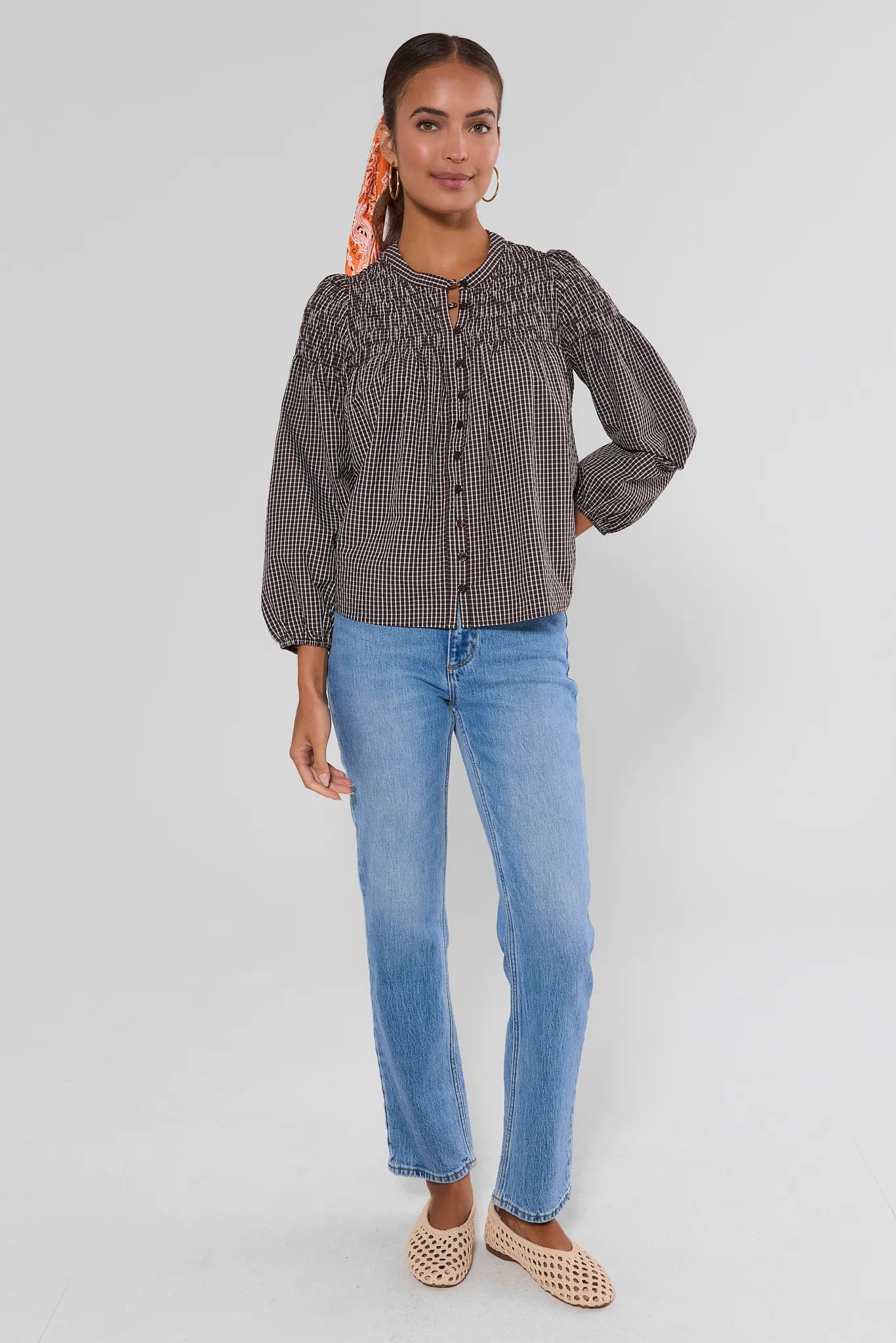 A woman modeling a dark gray and black micro-check patterned blouse with puff sleeves and a button-up front, paired with light blue jeans and beige woven flat shoes, against a light gray studio background.