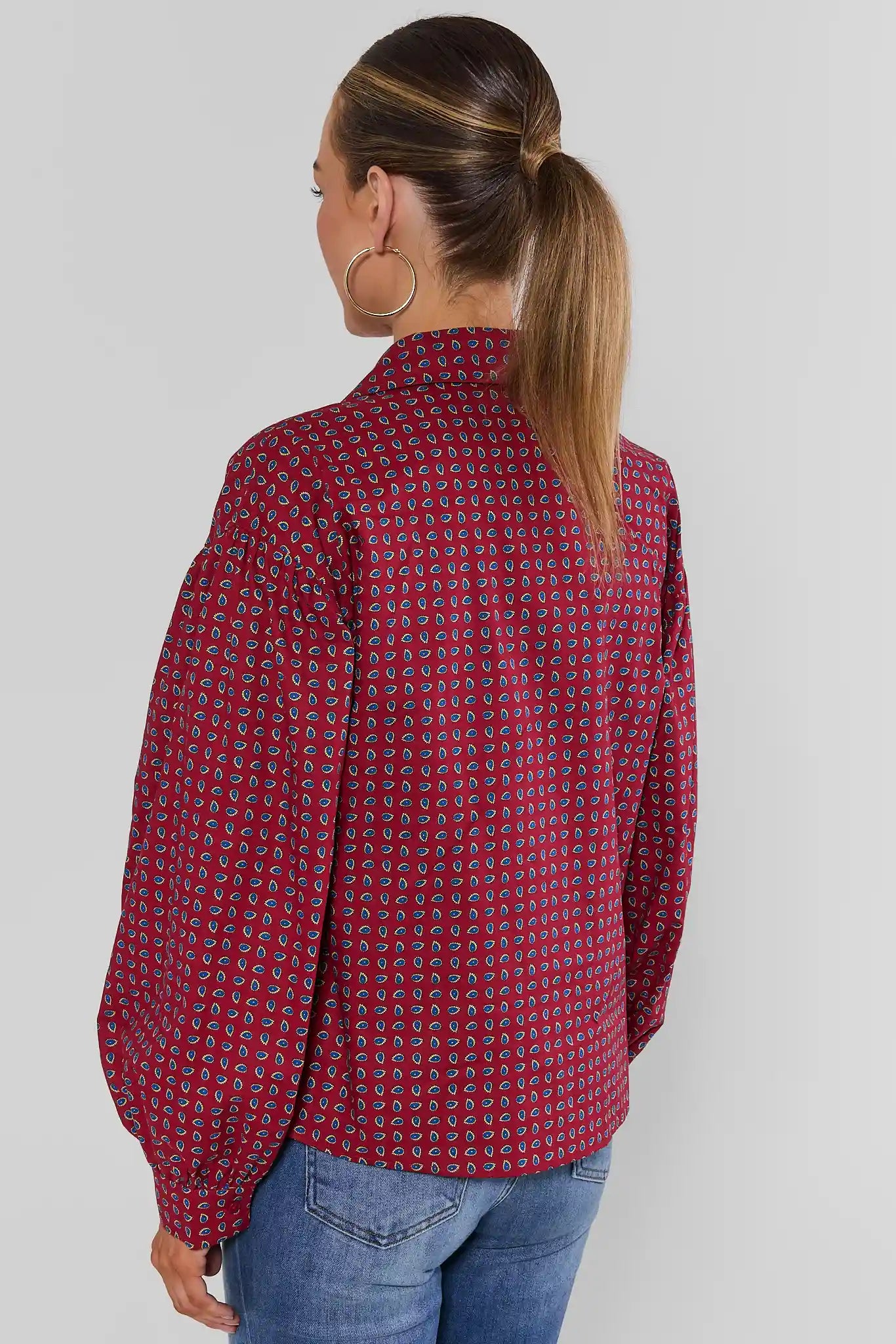A woman shown from the back wearing a red long-sleeve blouse with a dense small geometric pattern, paired with blue jeans, against a plain light gray studio background.