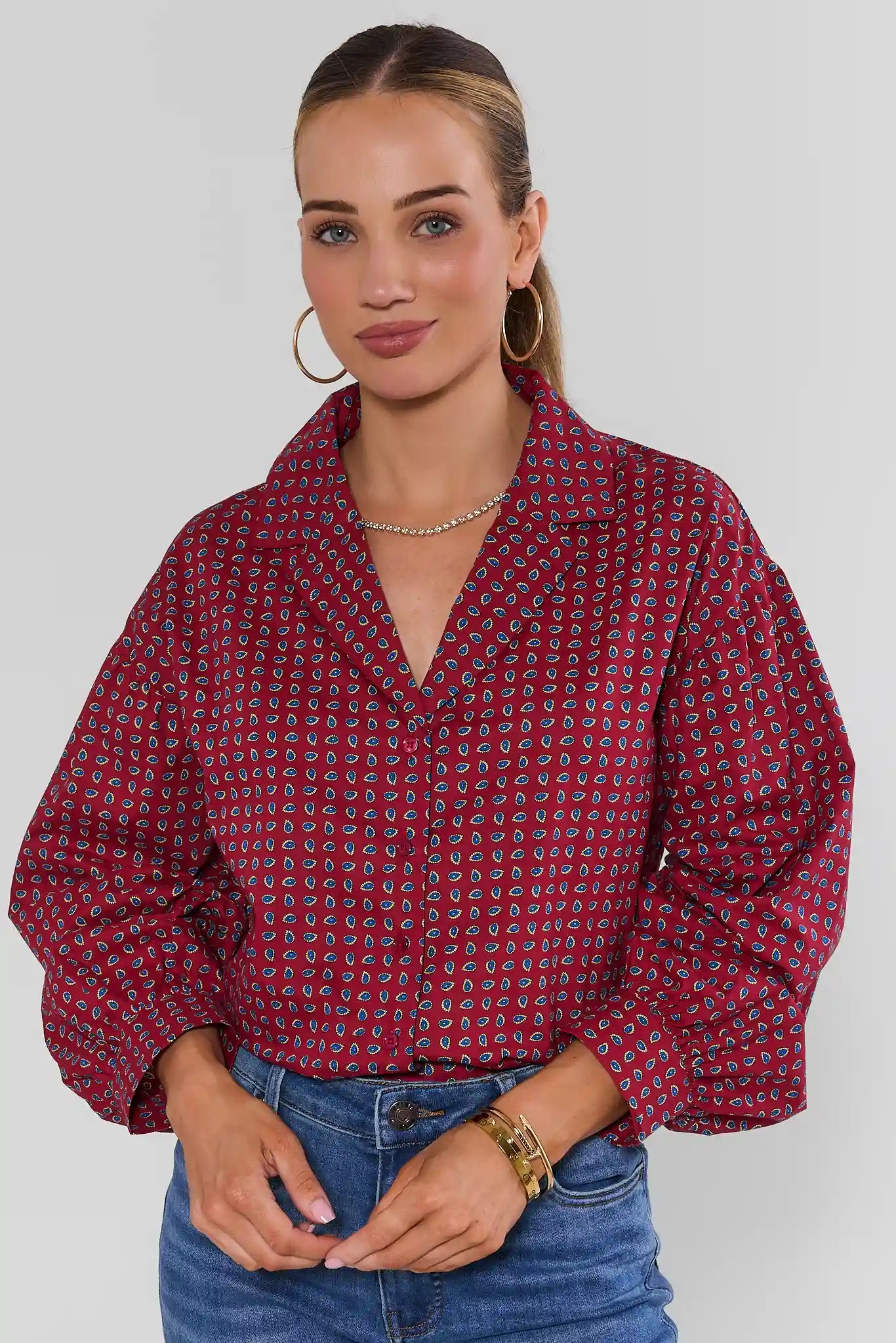 A woman wearing a red and blue patterned button-up blouse with voluminous puff sleeves, paired with blue jeans, against a light gray studio background.