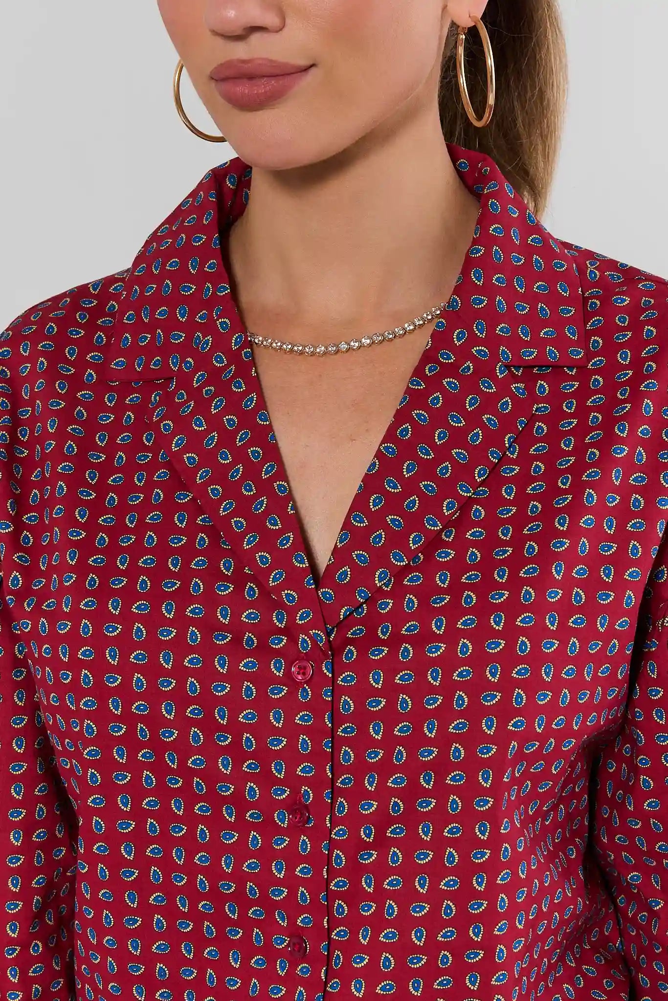 A close-up of a woman wearing a red collared button-up blouse with a blue and white paisley pattern, styled with gold hoop earrings and a delicate silver necklace.