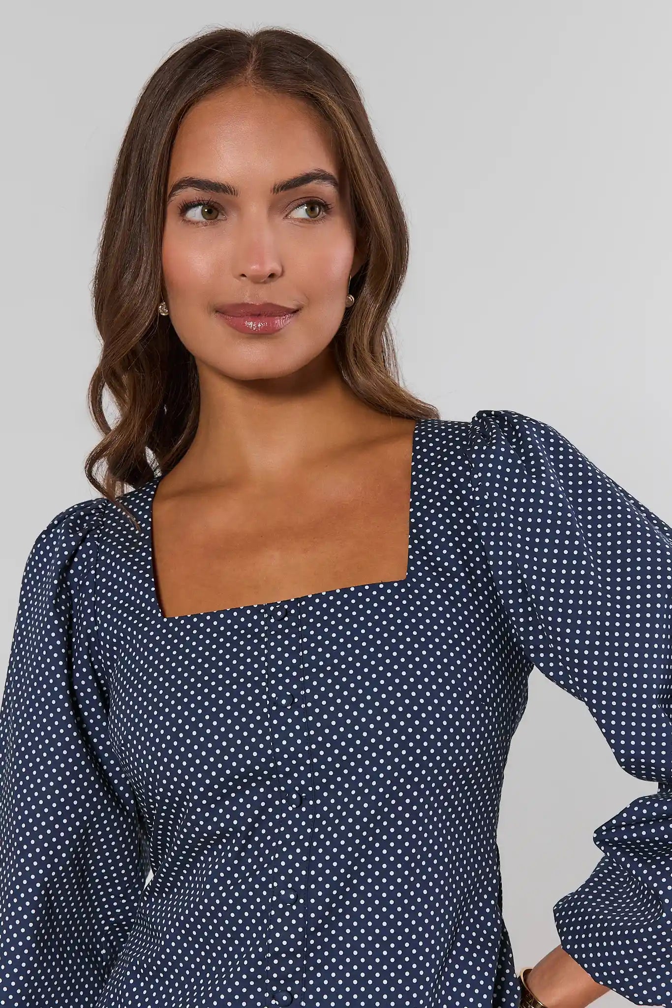A woman wearing a navy blue long-sleeve blouse with a white polka dot pattern and a square neckline, shown against a plain light gray studio background.