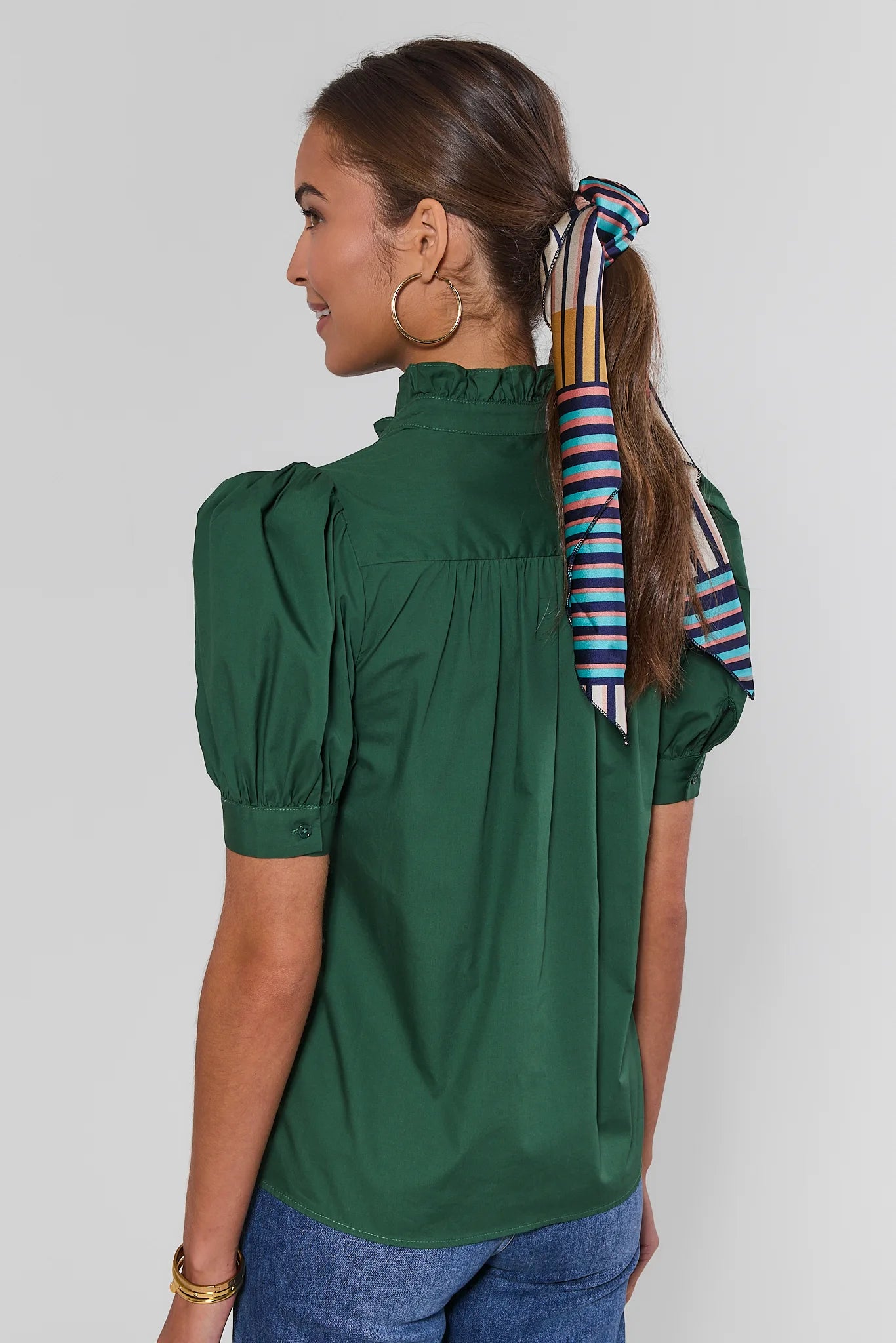 A woman shown from the back wearing a dark green short-sleeve blouse with puffed shoulders and a striped neck scarf tied in a ponytail, against a light gray studio background.