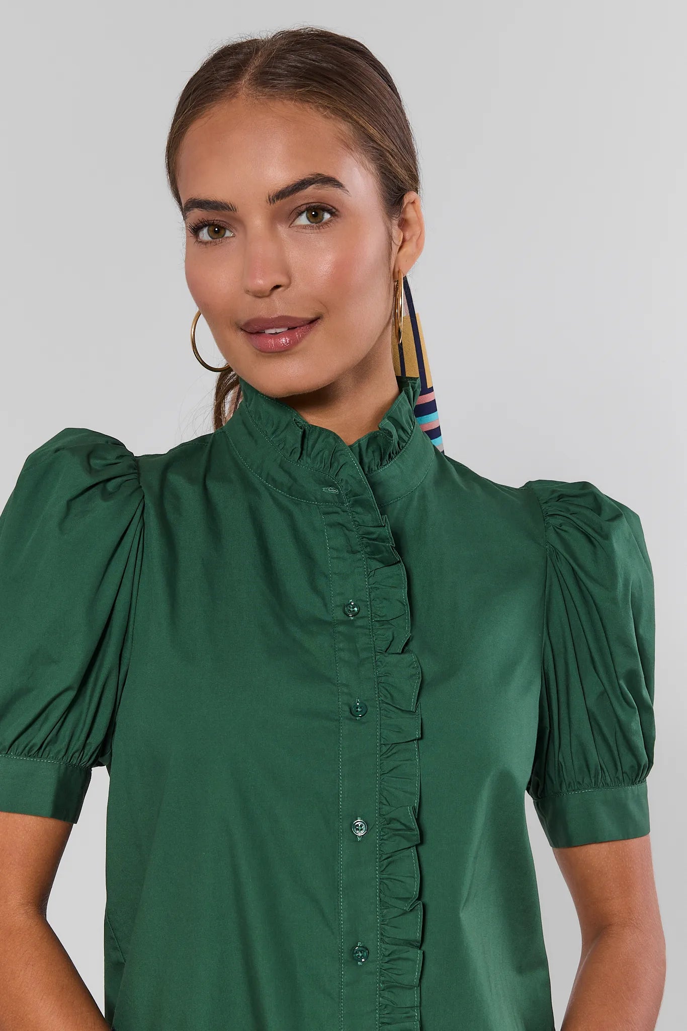 A woman wearing a dark green short-sleeve blouse with puffed sleeves, a ruffled high collar, and a button-front placket, photographed against a light studio background.