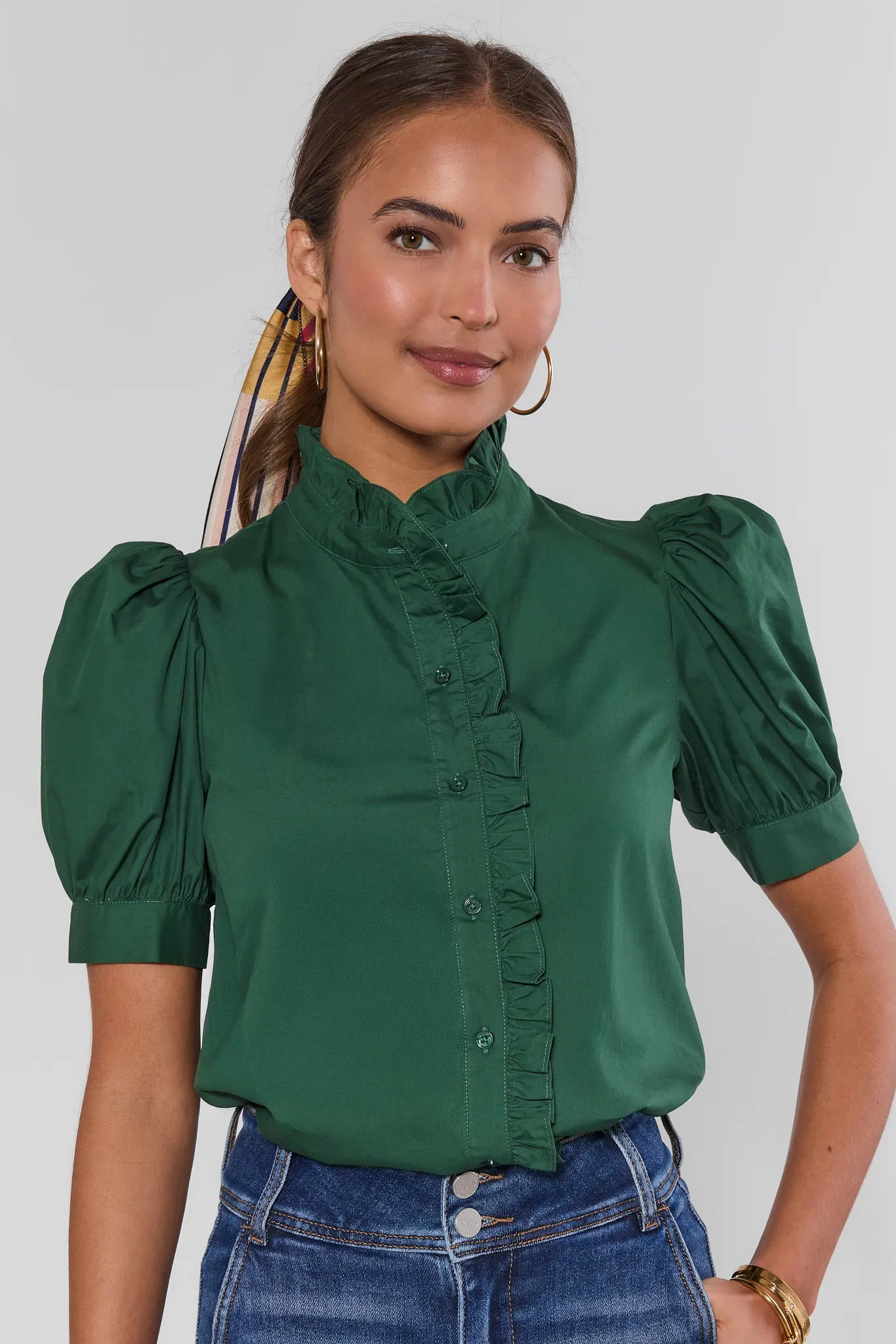 A woman wearing a dark green short-sleeve blouse with a high ruffled collar and puffed sleeves, paired with blue jeans, against a light gray studio background.