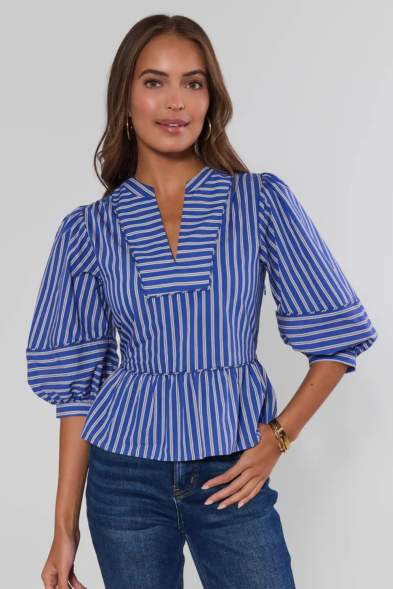 A woman wearing a blue and white vertically striped blouse with puff sleeves and a peplum waist, paired with blue jeans, against a light gray studio background.
