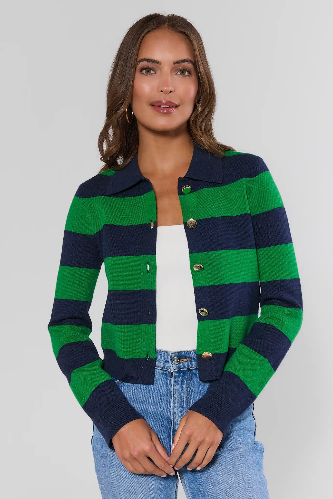 A woman wearing a green and navy blue horizontally striped knit cardigan with gold buttons, styled over a white top and light wash blue jeans, against a plain light gray studio background.