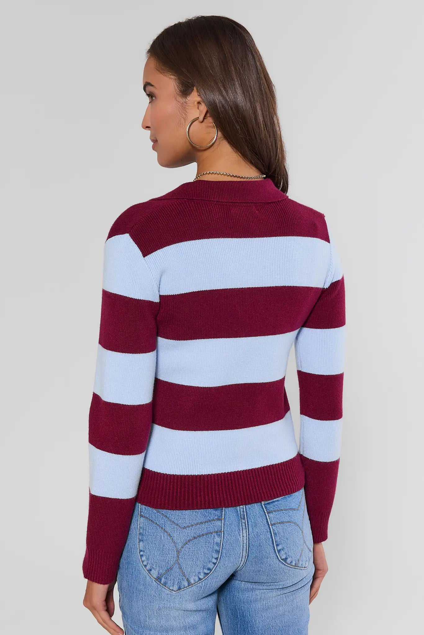 A woman shown from the back wearing a long-sleeve knit sweater with horizontal burgundy and light blue stripes, paired with light-wash blue jeans, against a plain light gray studio background.
