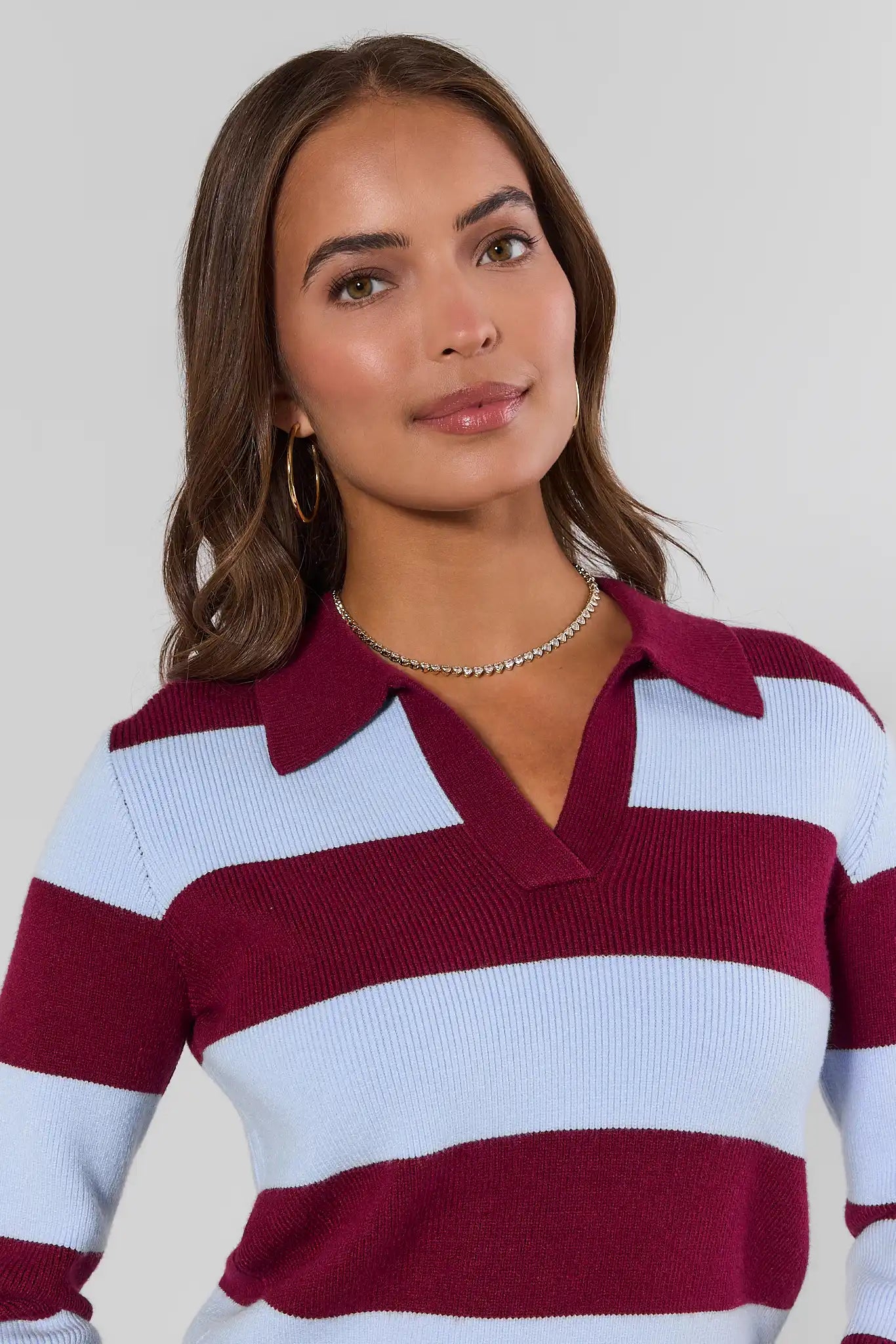 A woman wearing a ribbed knit polo sweater with horizontal stripes in deep burgundy and light blue, paired with a delicate silver chain necklace against a light gray background.
