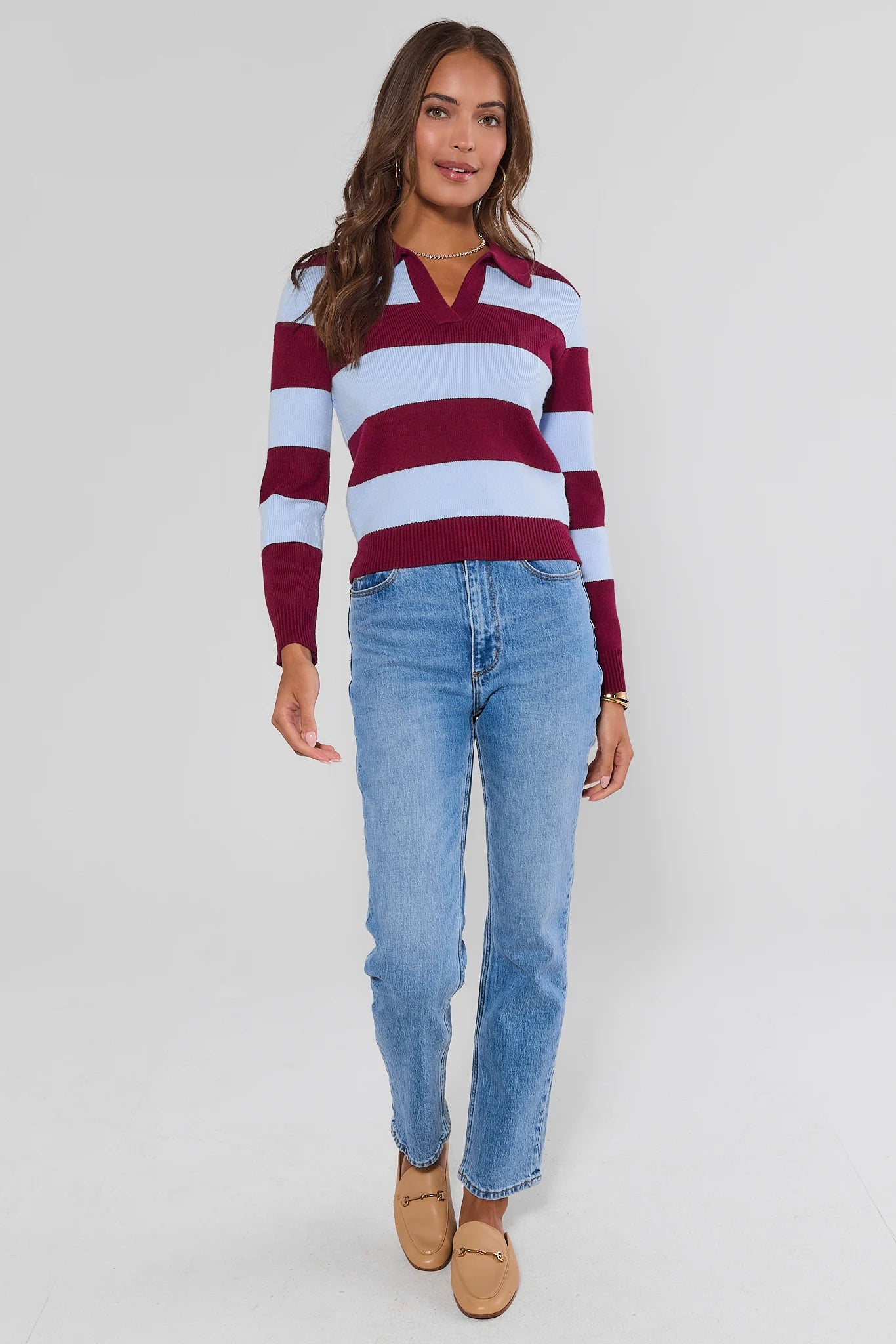 A woman modeling a long-sleeve knit polo sweater with horizontal burgundy and white stripes, paired with light-wash blue jeans and tan loafers, against a plain gray studio background.