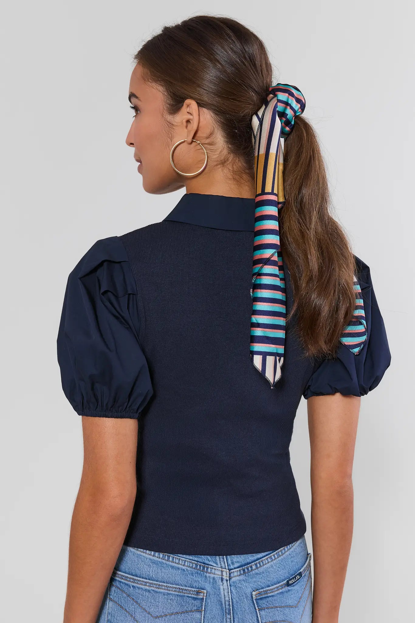A woman shown from the back wearing a navy blue puff-sleeve top with a colorful striped neck scarf tied in a ponytail, paired with blue jeans against a light gray studio background.