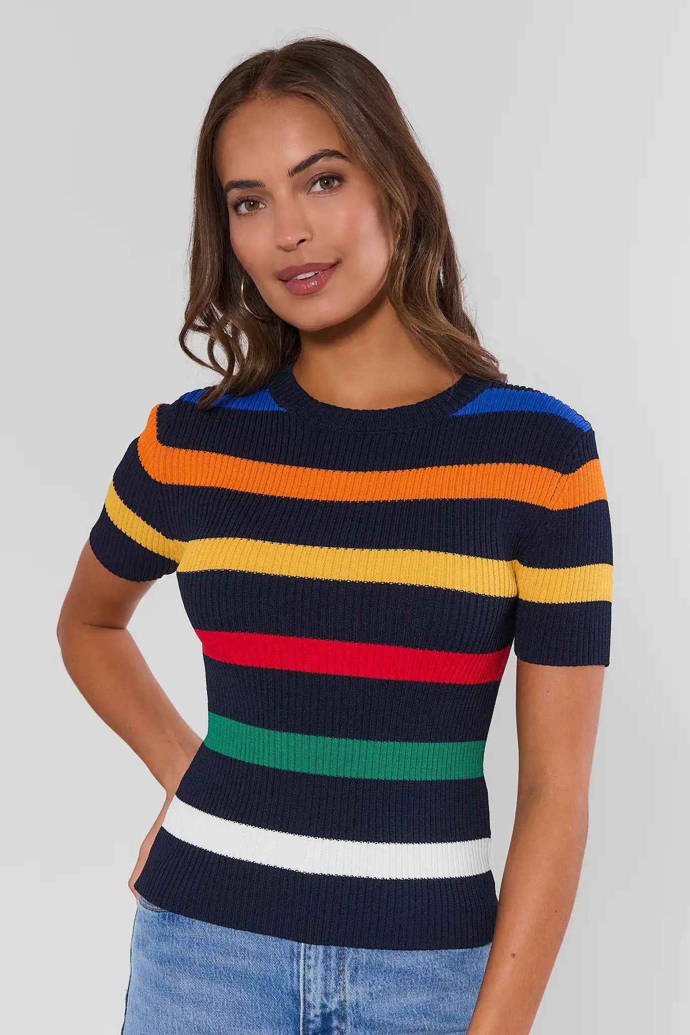 A woman wearing a short-sleeve ribbed knit sweater with horizontal stripes in navy, blue, orange, yellow, red, green, and white, paired with light blue jeans, against a light gray studio background.