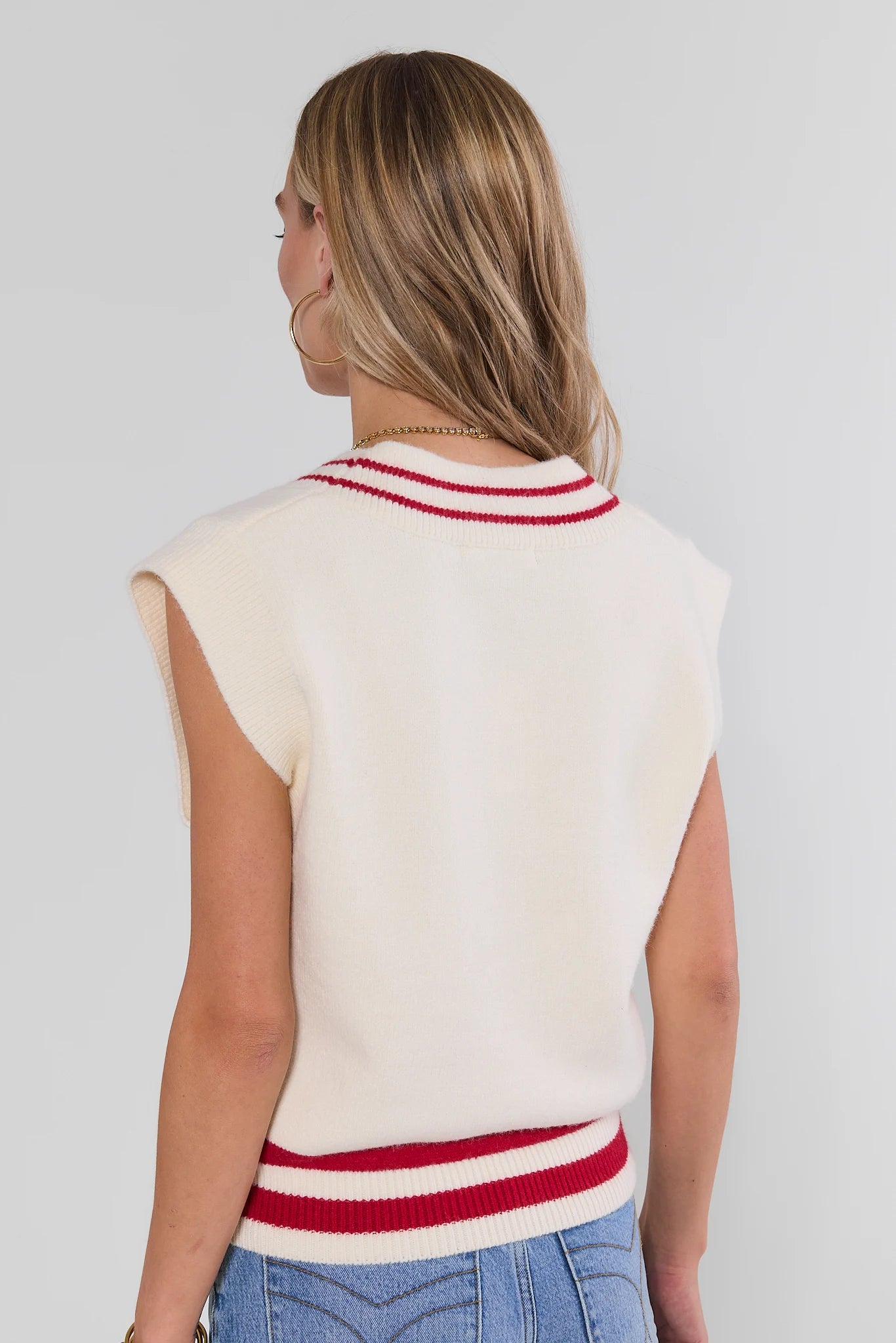 A woman shown from the back wearing a sleeveless cream knit sweater vest with red and white striped trim at the collar and hem, paired with blue jeans against a light gray studio background.
