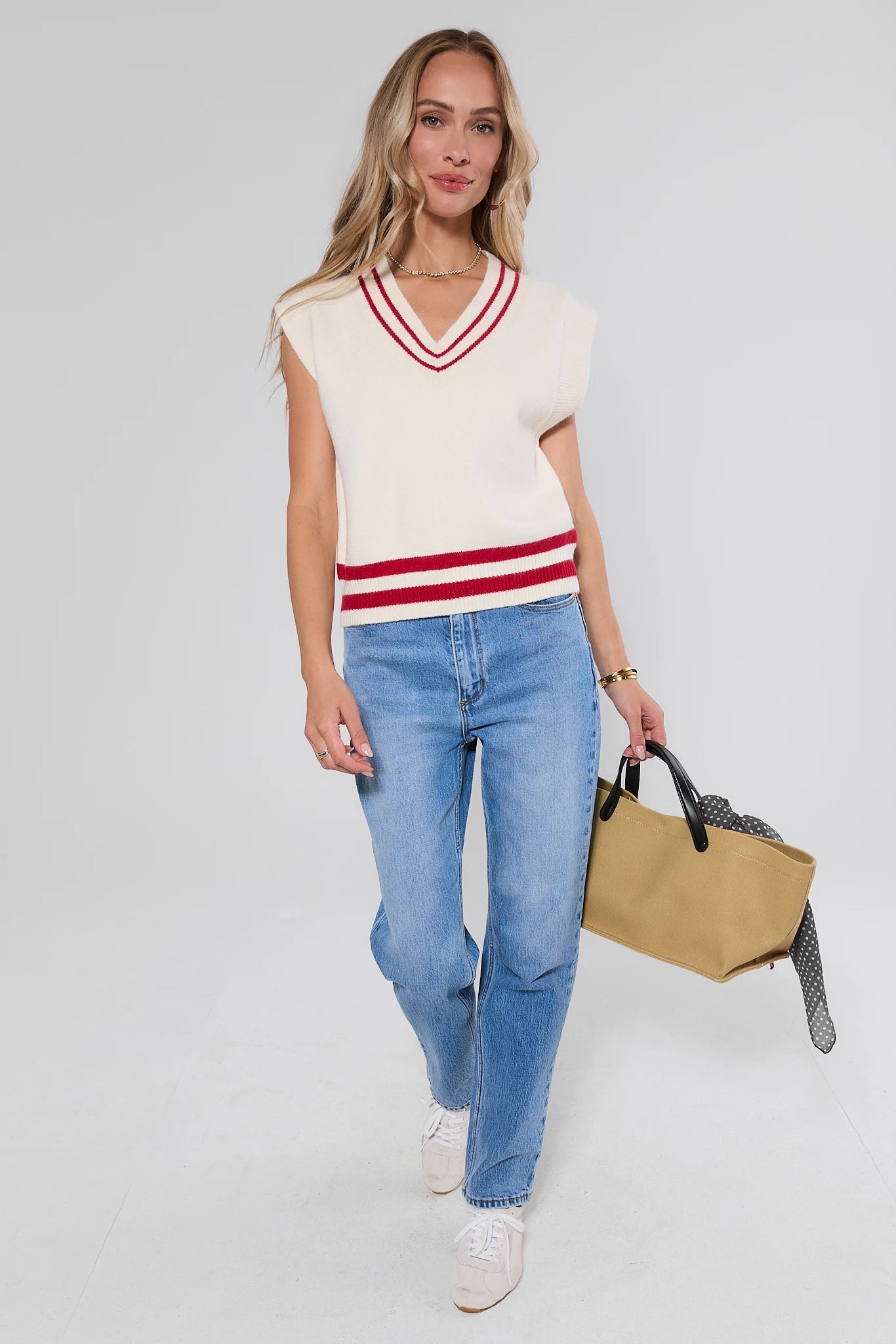 A woman modeling a cream knit V-neck sweater vest with red stripes, paired with light blue jeans, white sneakers, and holding a tan tote bag against a light gray studio background.