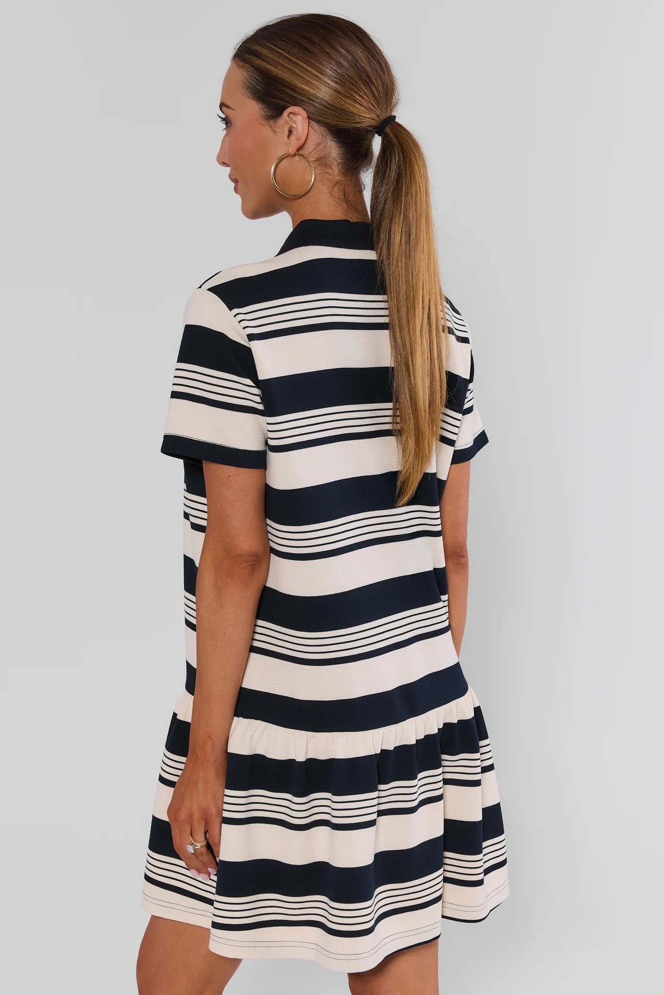 A woman wearing a short-sleeve striped dress, shown from the back. The dress has horizontal navy blue and cream stripes, and is photographed against a plain light gray studio background.