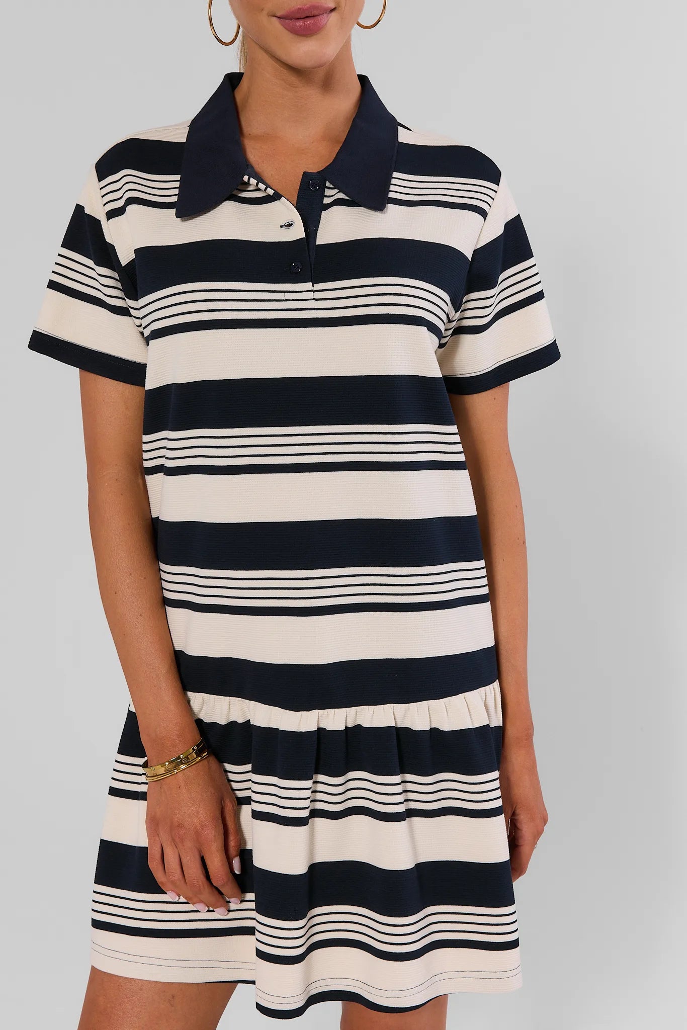 A woman wearing a short-sleeve polo dress with horizontal navy blue and cream stripes, featuring a navy collar and a buttoned placket, set against a plain light gray background.
