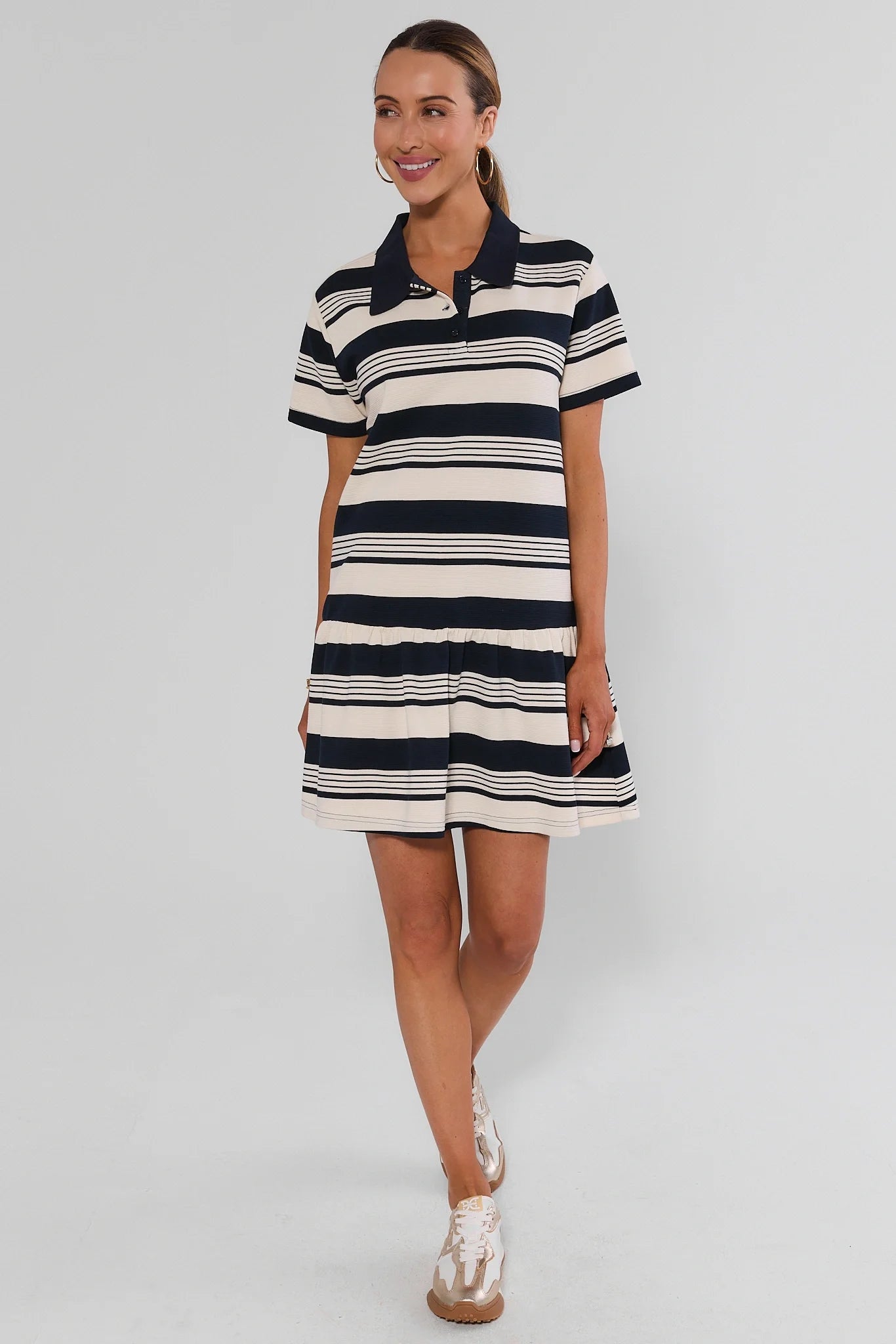 A woman modeling a short-sleeve polo dress with horizontal navy and cream stripes, paired with white sneakers, against a light gray background.