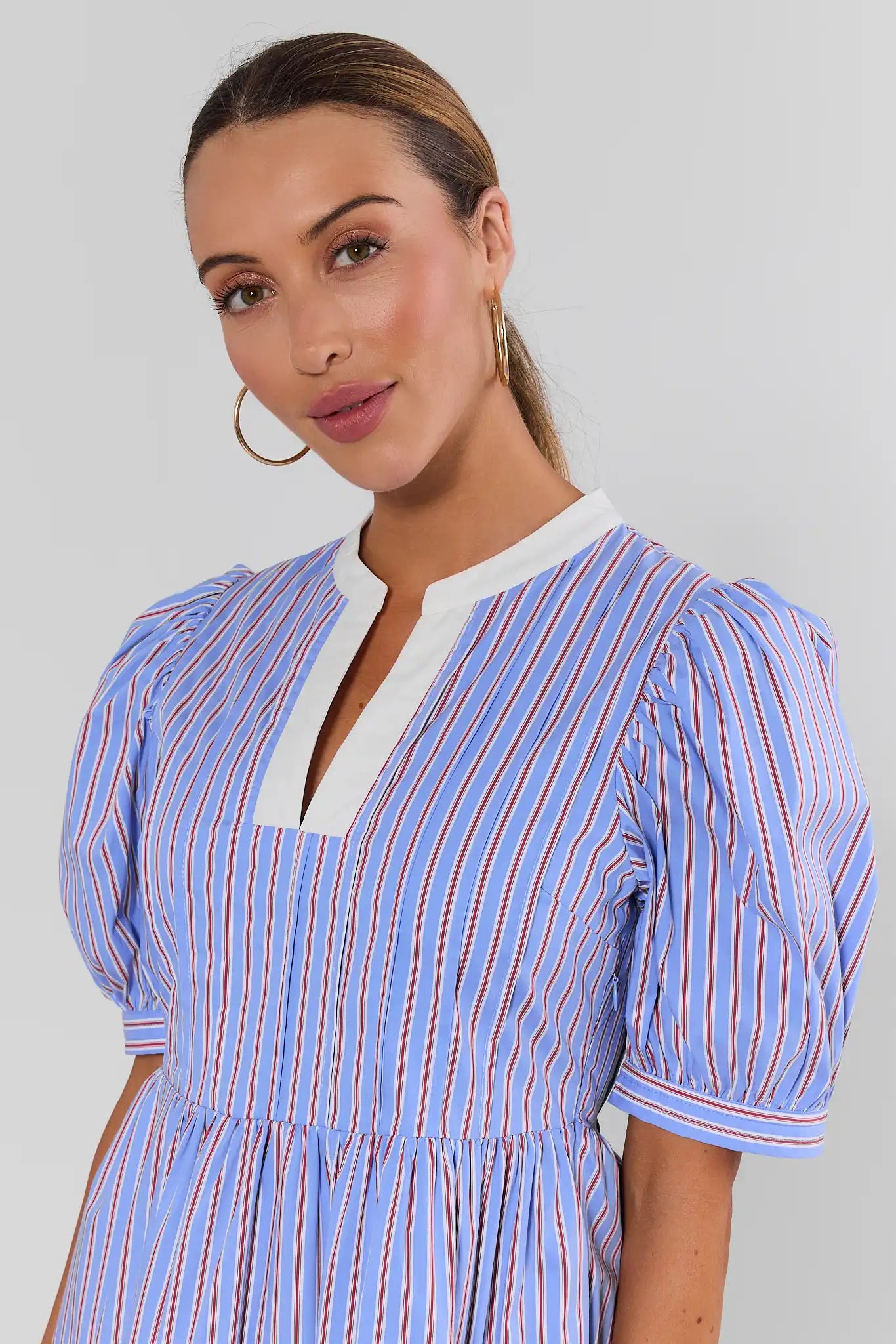 A woman wearing a light blue and white vertically striped puff-sleeve dress with a white split neckline, photographed against a plain light gray studio background.