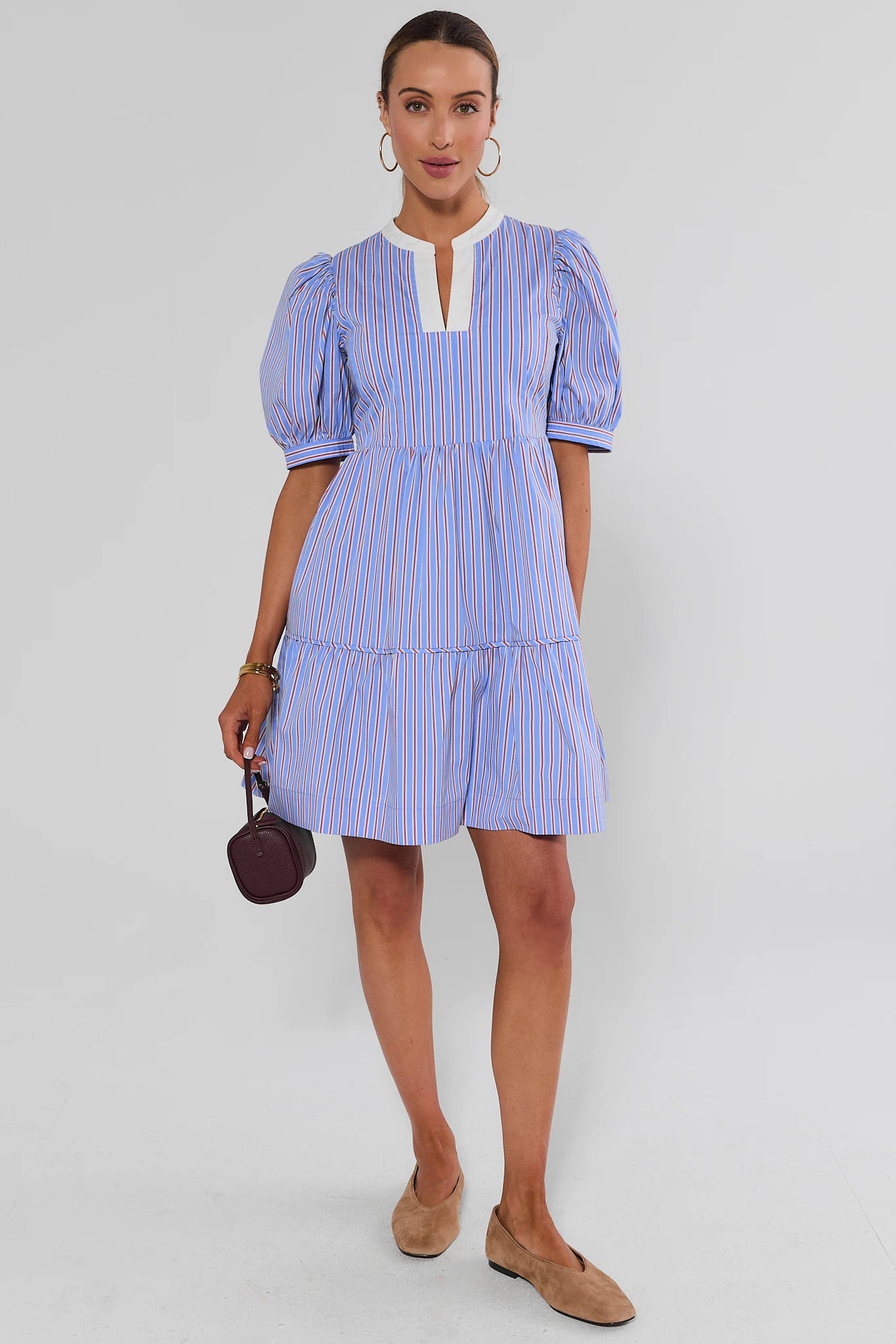 A woman modeling a light blue and white vertically striped puff-sleeve mini dress with a white keyhole neckline, paired with tan flat shoes and holding a small dark handbag, against a light gray studio background.