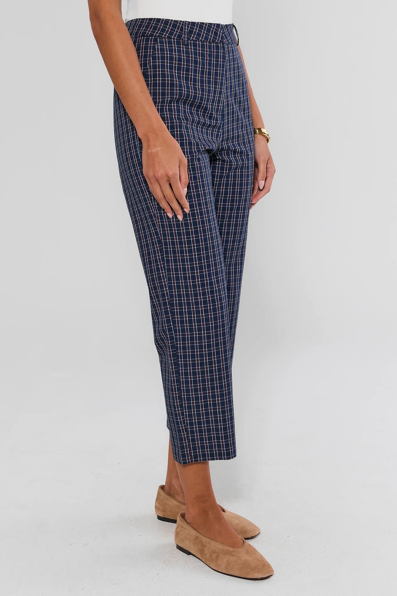 A person wearing navy blue plaid cropped trousers with a white top and tan suede flats, shown from the waist down against a light gray studio background.
