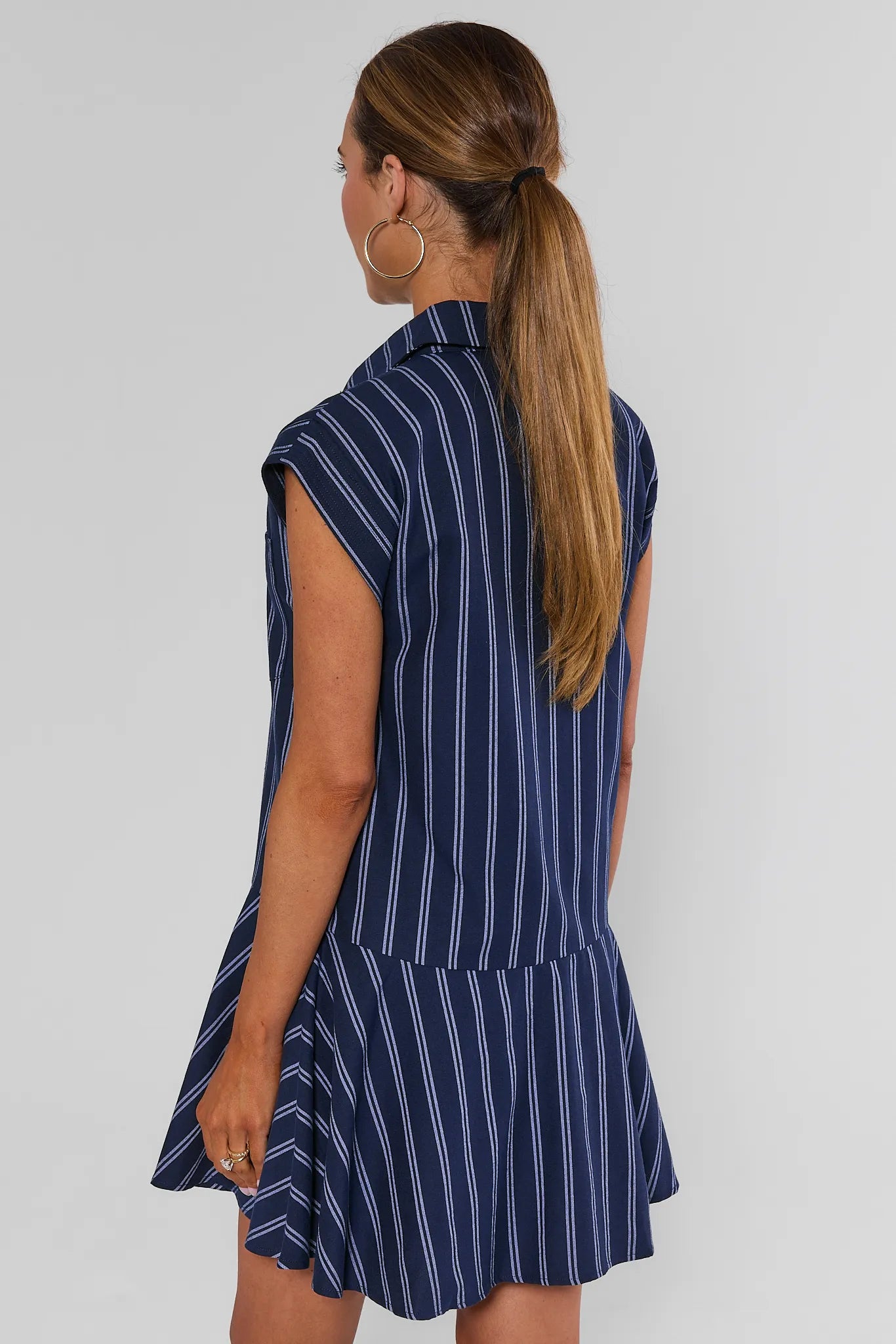 A woman shown from the back wearing a sleeveless navy blue dress with thin white vertical pinstripes and a flared ruffled hem, against a plain light gray studio background.