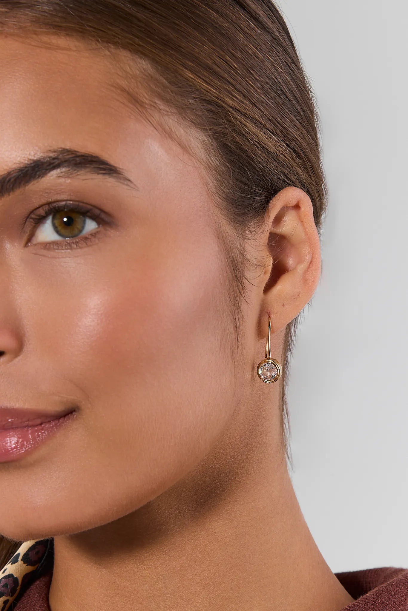 A close-up of a woman's ear wearing a delicate gold dangle earring with a round clear gemstone, set against a neutral light gray background.